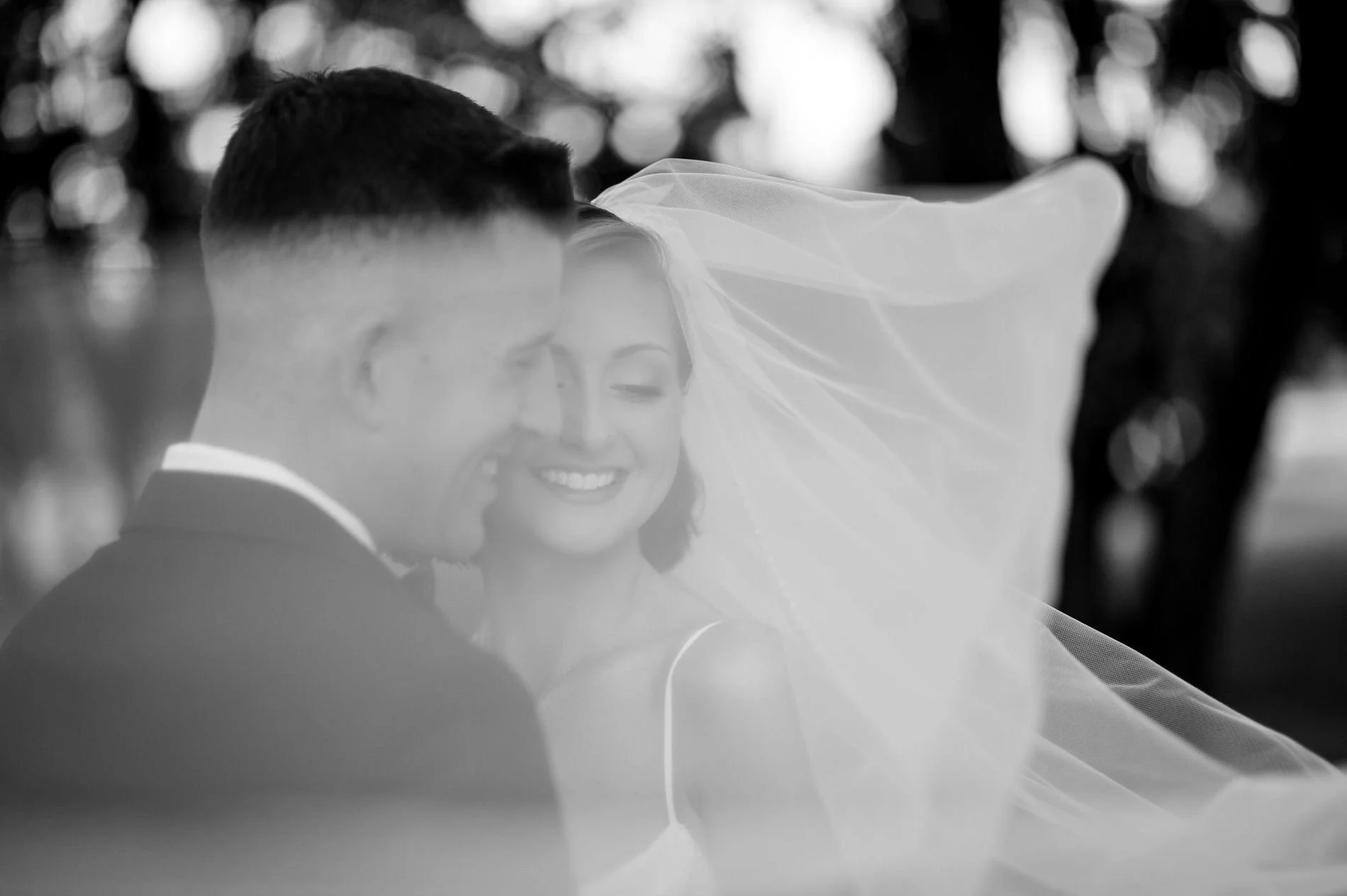Black and white photo of Emily and Austin cheek to cheek as her veil blows around them