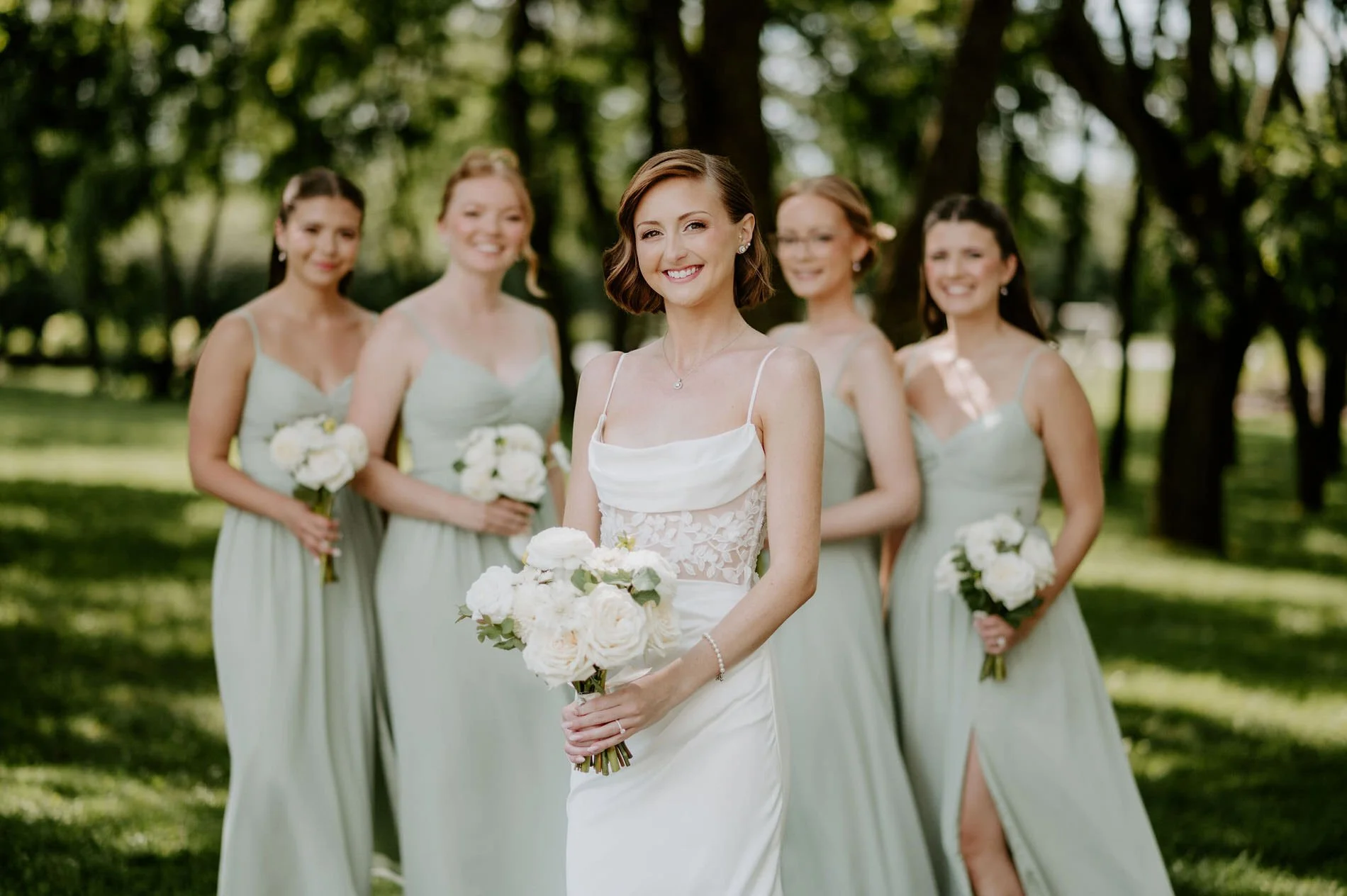 Emily holding her white bouquet with her bridesmaids in sage gowns behind her on the lawn at Stonefields Estate