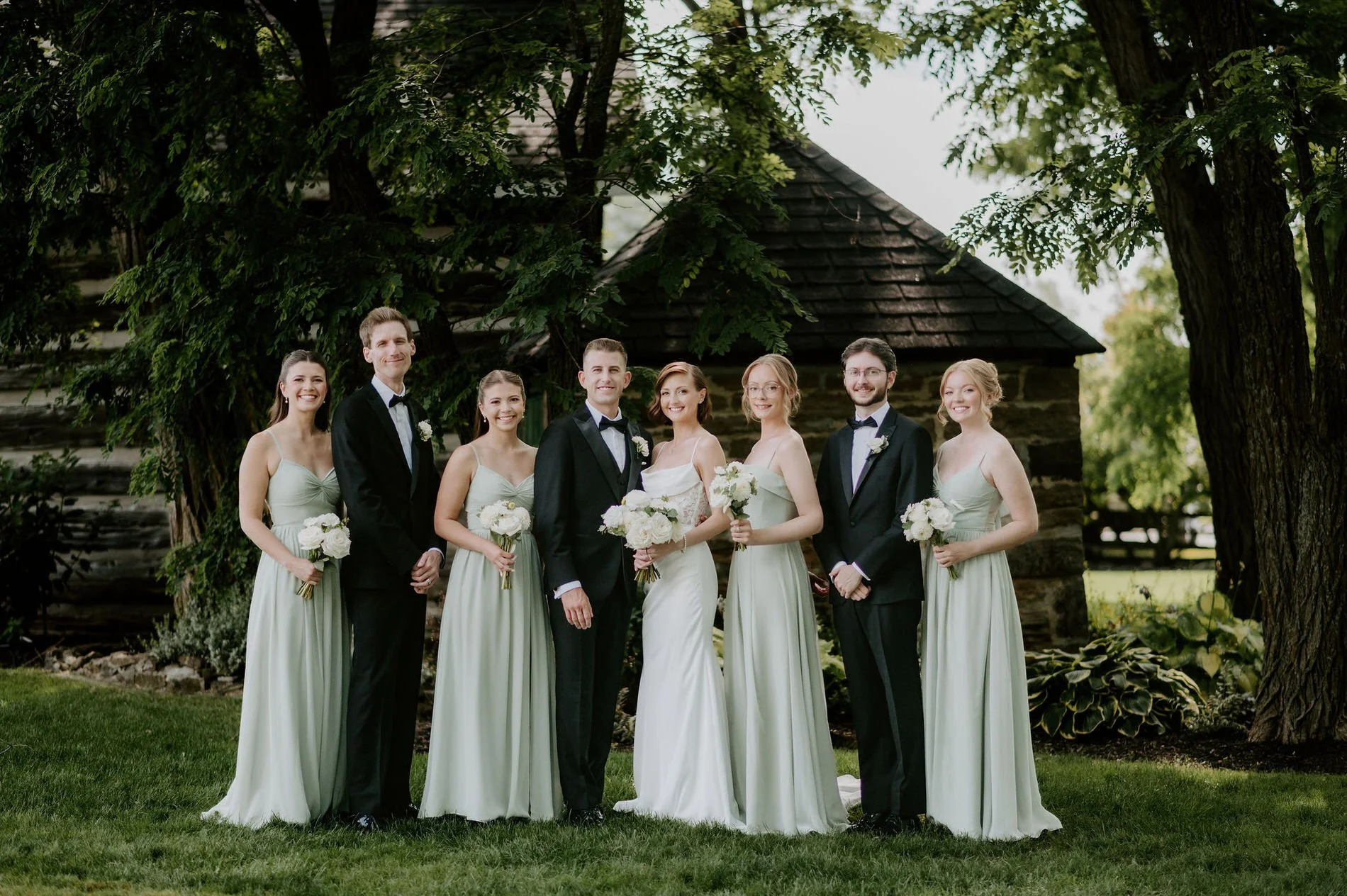 Emily and Austin's wedding party in sage gowns and black tuxedos in front of a log building at Stonefields Estate