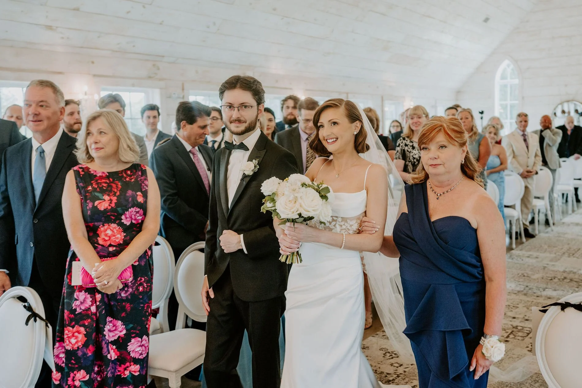 Emily walking down the chapel aisle between a young man in a tuxedo and a woman in a navy gown