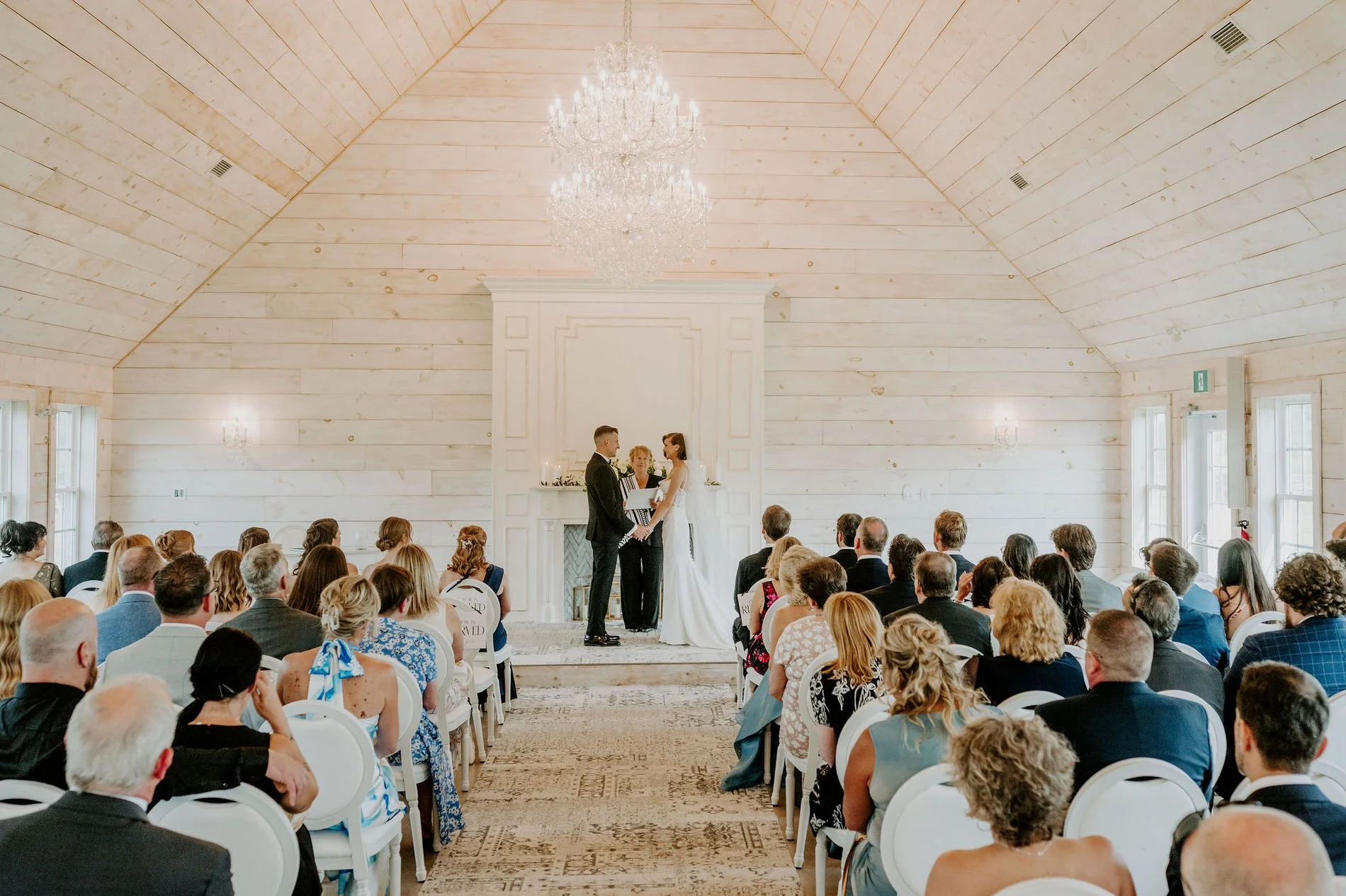 Wide view of Emily and Austin's ceremony inside the white chapel at Stonefields Estate, under a crystal chandelier
