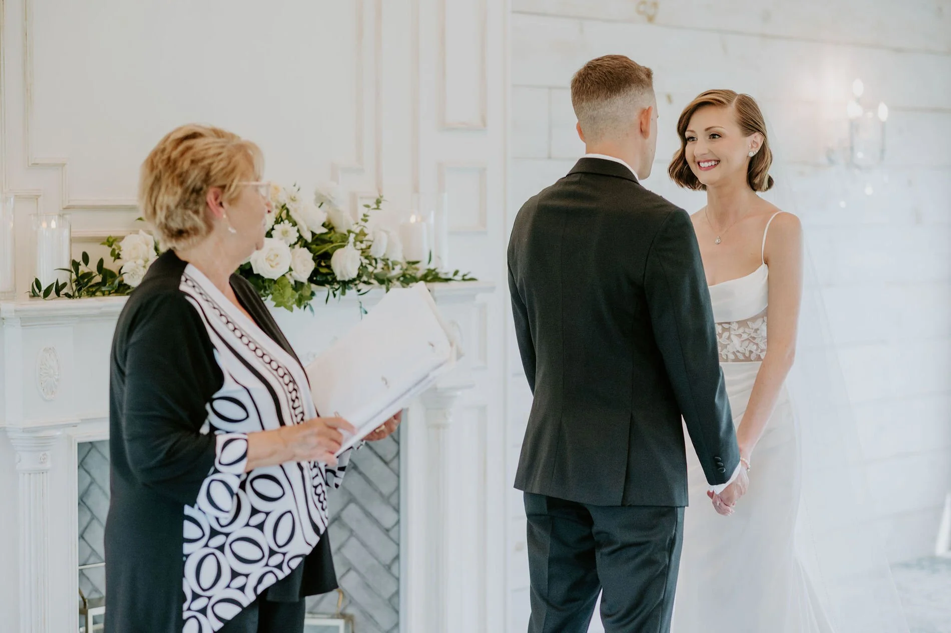 Emily smiling as she holds Austin's hands while the officiant reads beside a white fireplace