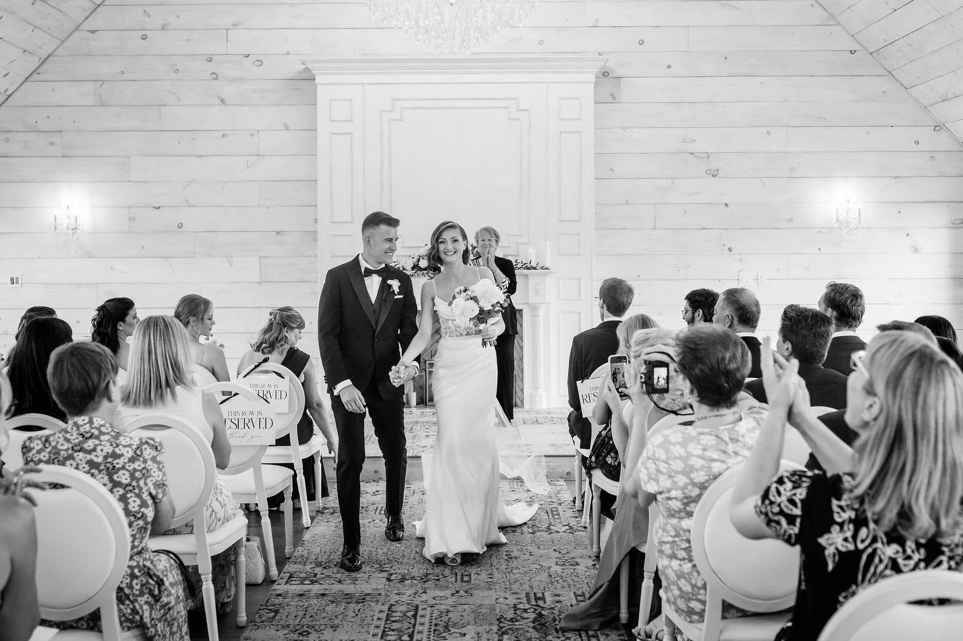Black and white photo of Emily and Austin walking back up the chapel aisle as guests take photos