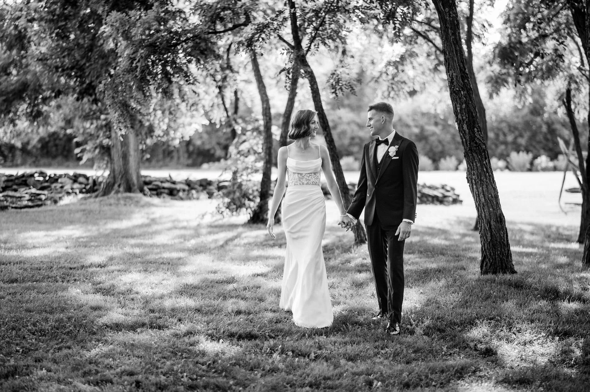 Black and white photo of Emily and Austin walking hand in hand beneath the trees at Stonefields Estate