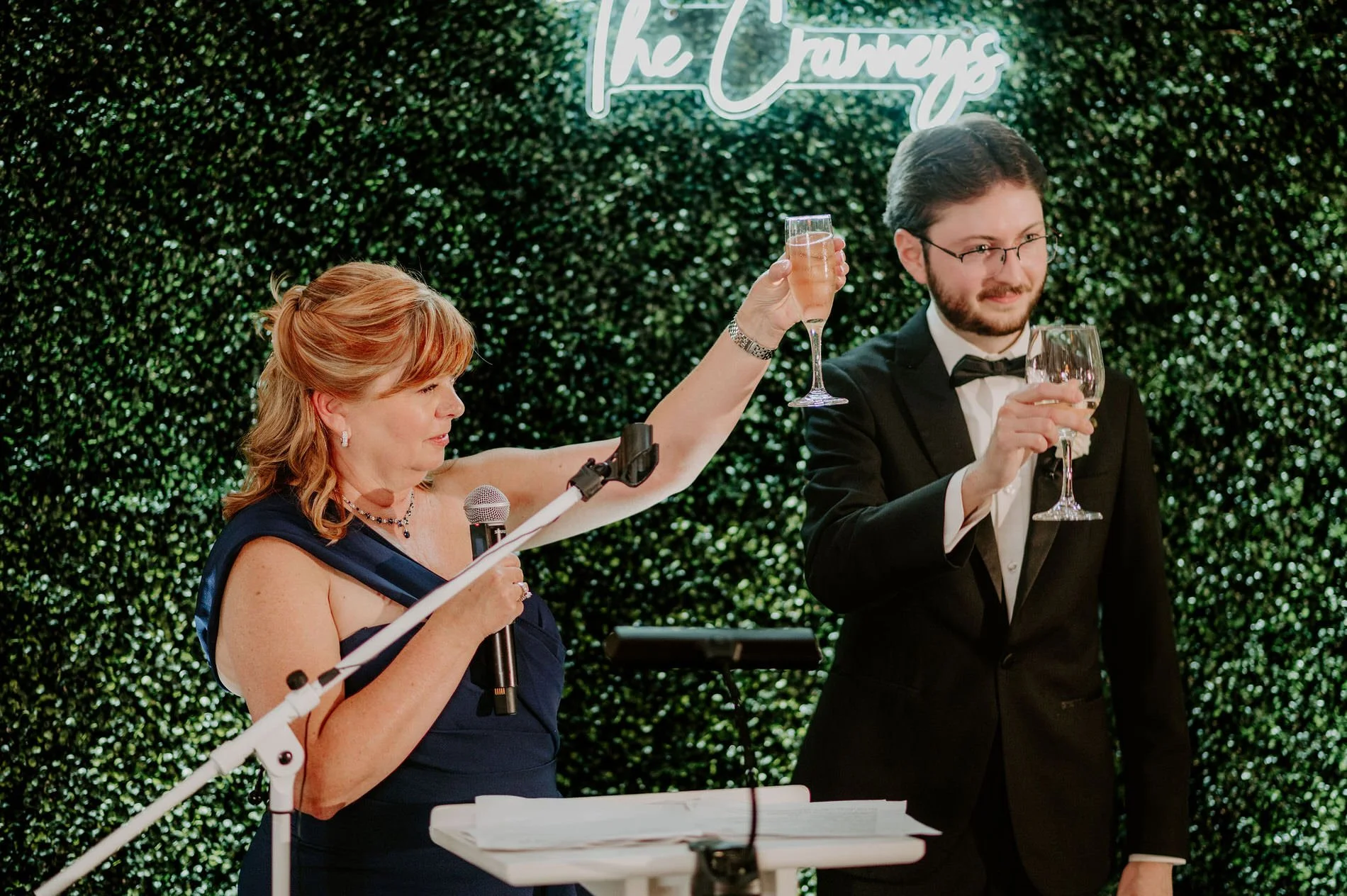 A woman in a navy gown and a young man in a tuxedo raising their glasses for a toast in front of a hedge wall