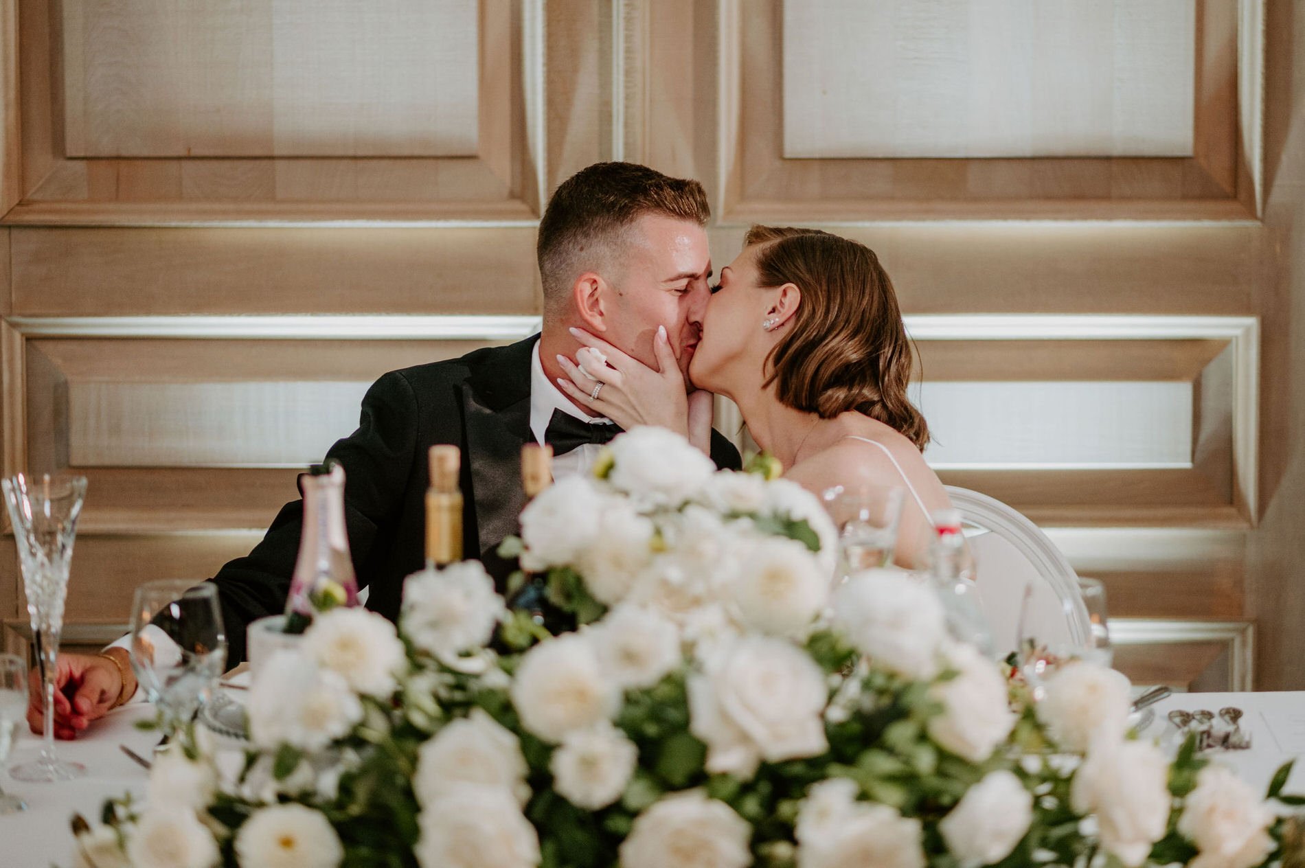 Emily and Austin sharing a kiss at the head table behind white roses