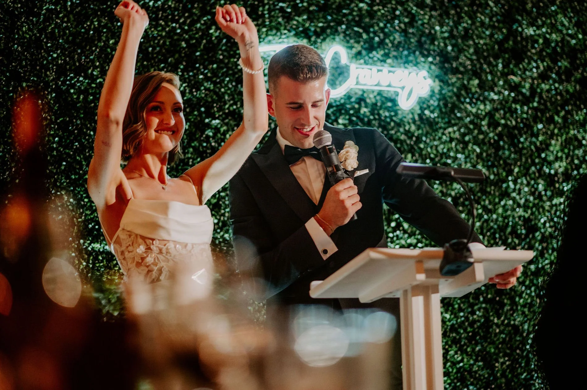 Emily raising her arms in celebration as Austin gives a speech in front of a neon-lit hedge wall