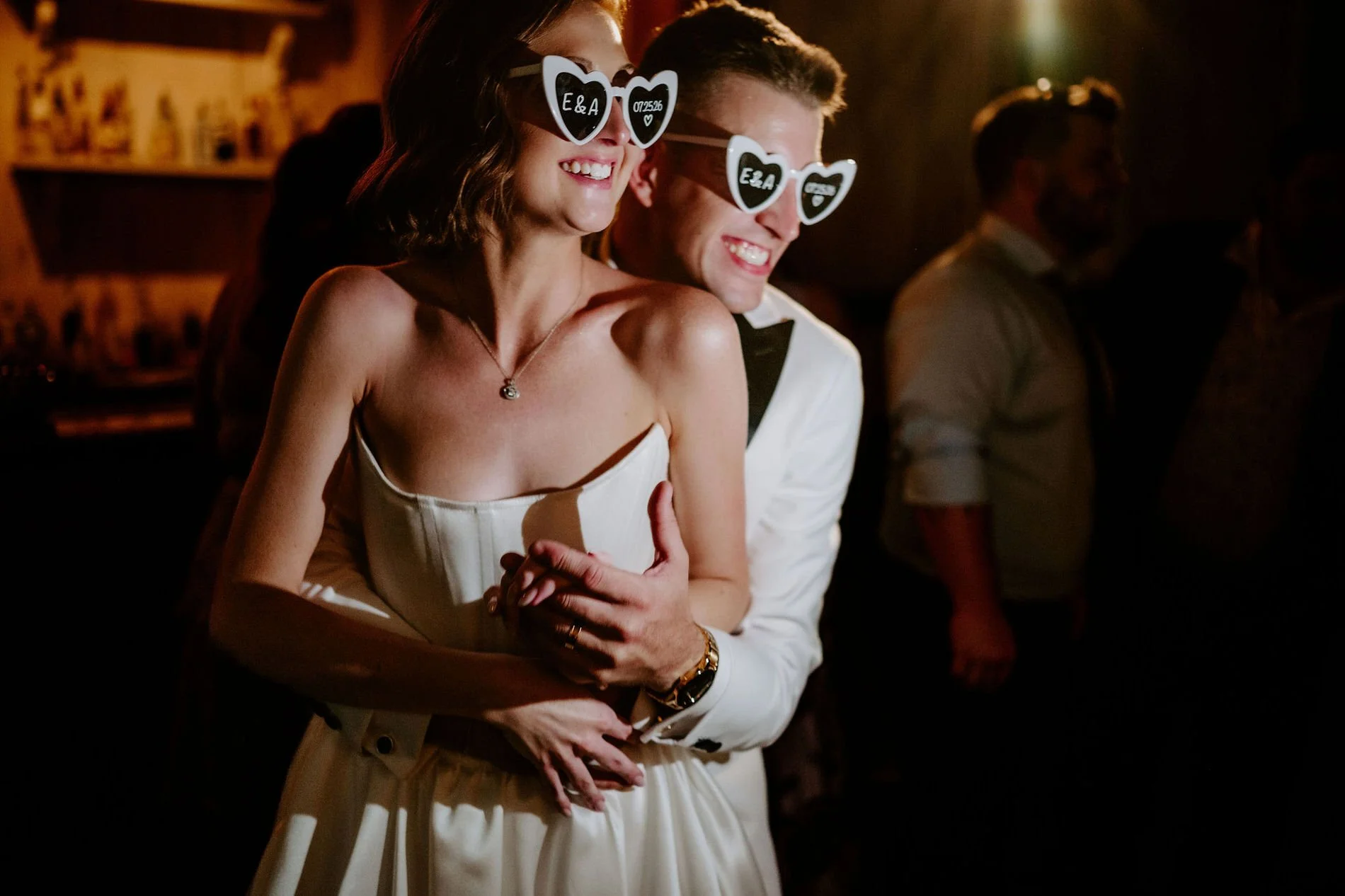 Austin hugging Emily from behind, both laughing in heart-shaped "E&amp;A" sunglasses
