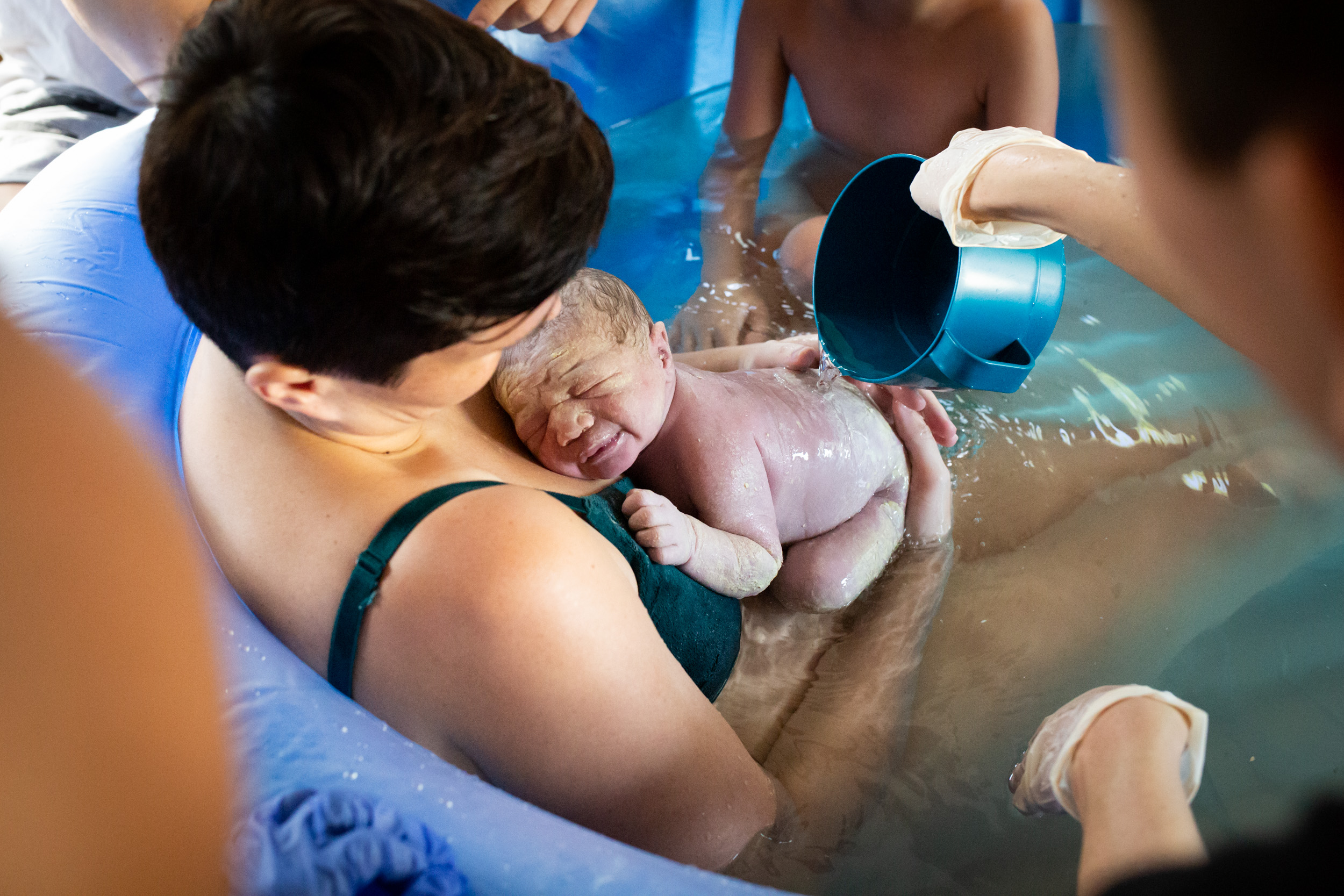 mom holding newborn baby in birthing tub as midwife pours warm water on them