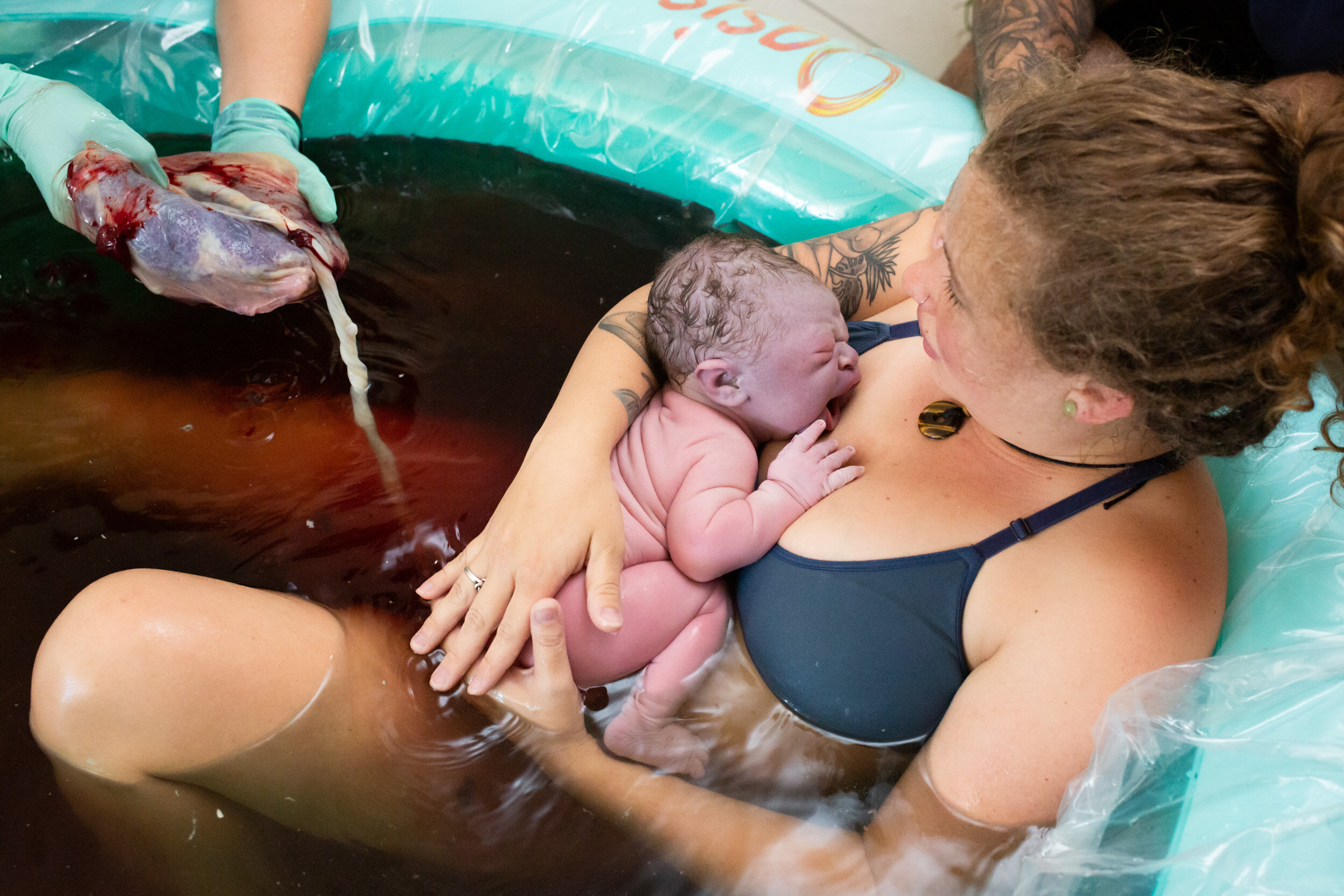 mom holding newborn baby in birthing pool while the midwife holds the placenta