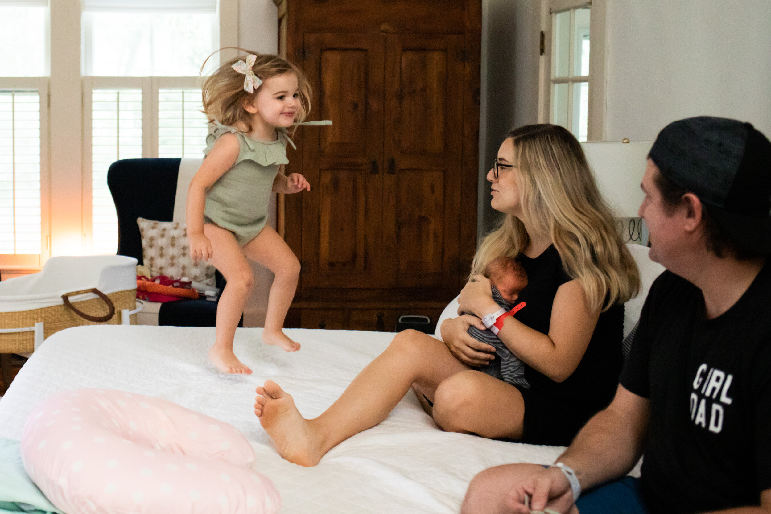 girl jumping on bed with family and newborn baby sister being held by mom