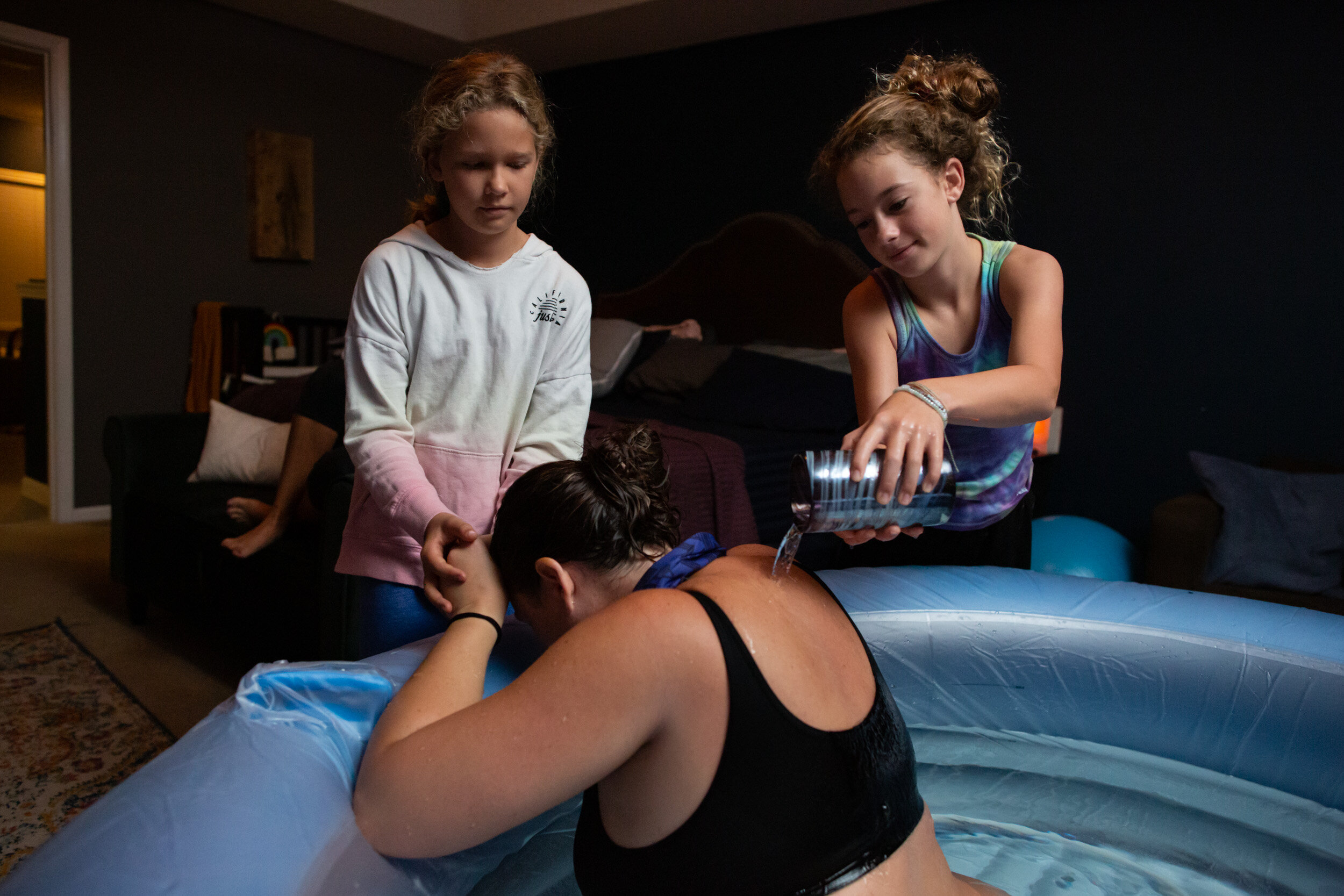 daughters pouring water on their mom's back while she labors in birth tub