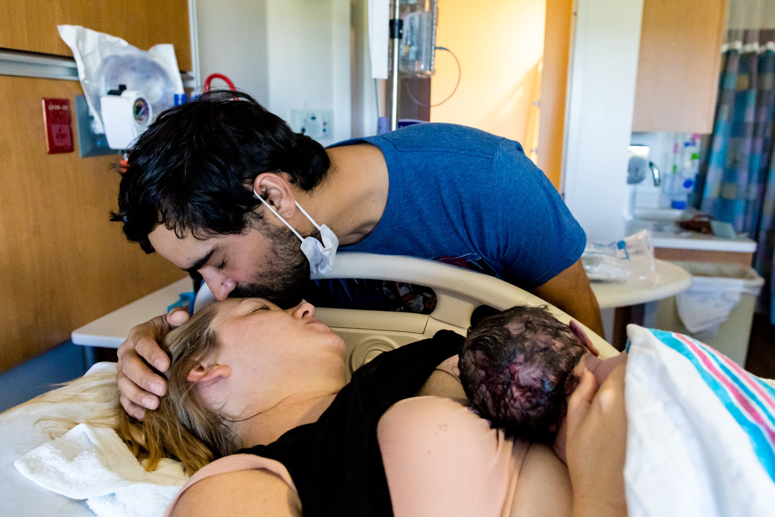 husband kissing his wife's forehead as she holds their newborn baby