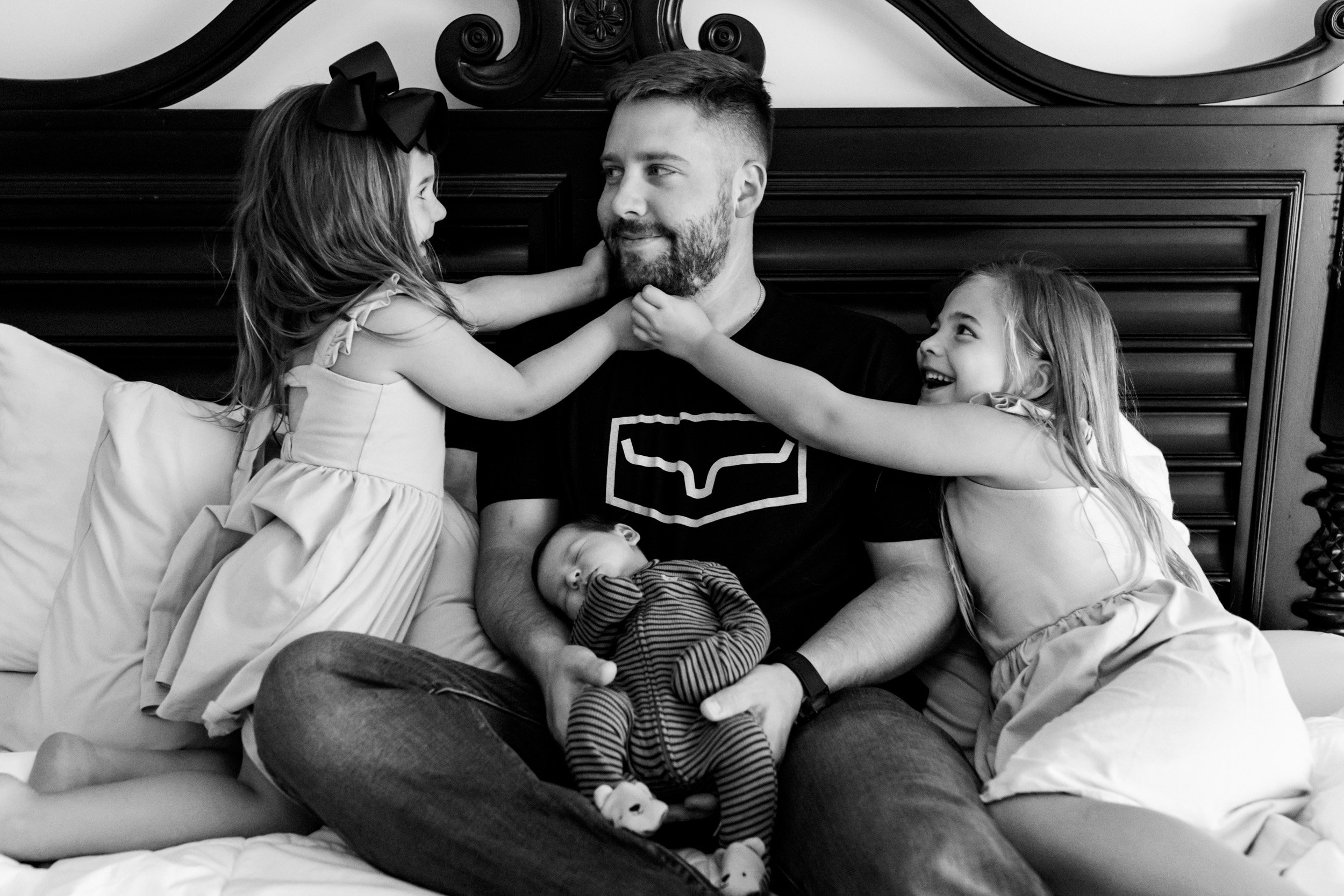 dad holding newborn baby while his daughters tickle his face
