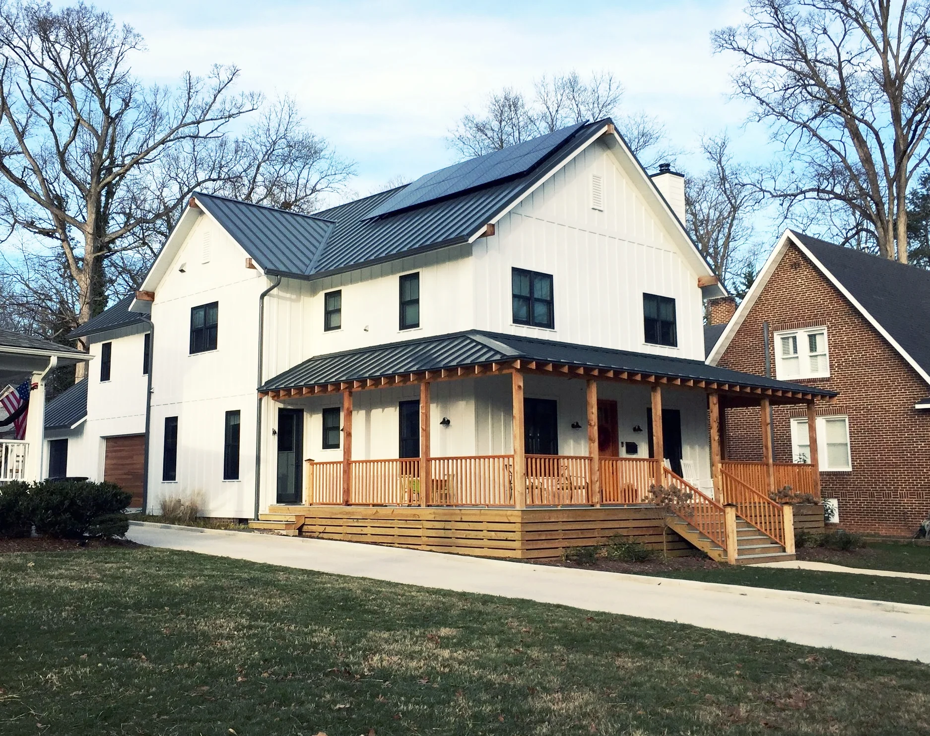 A two-story house with a white exterior and black windows, featuring a metal roof and a wooden porch, next to a brick house with a steep roof. The yard has green grass and a concrete walkway, with leafless trees in the background.
