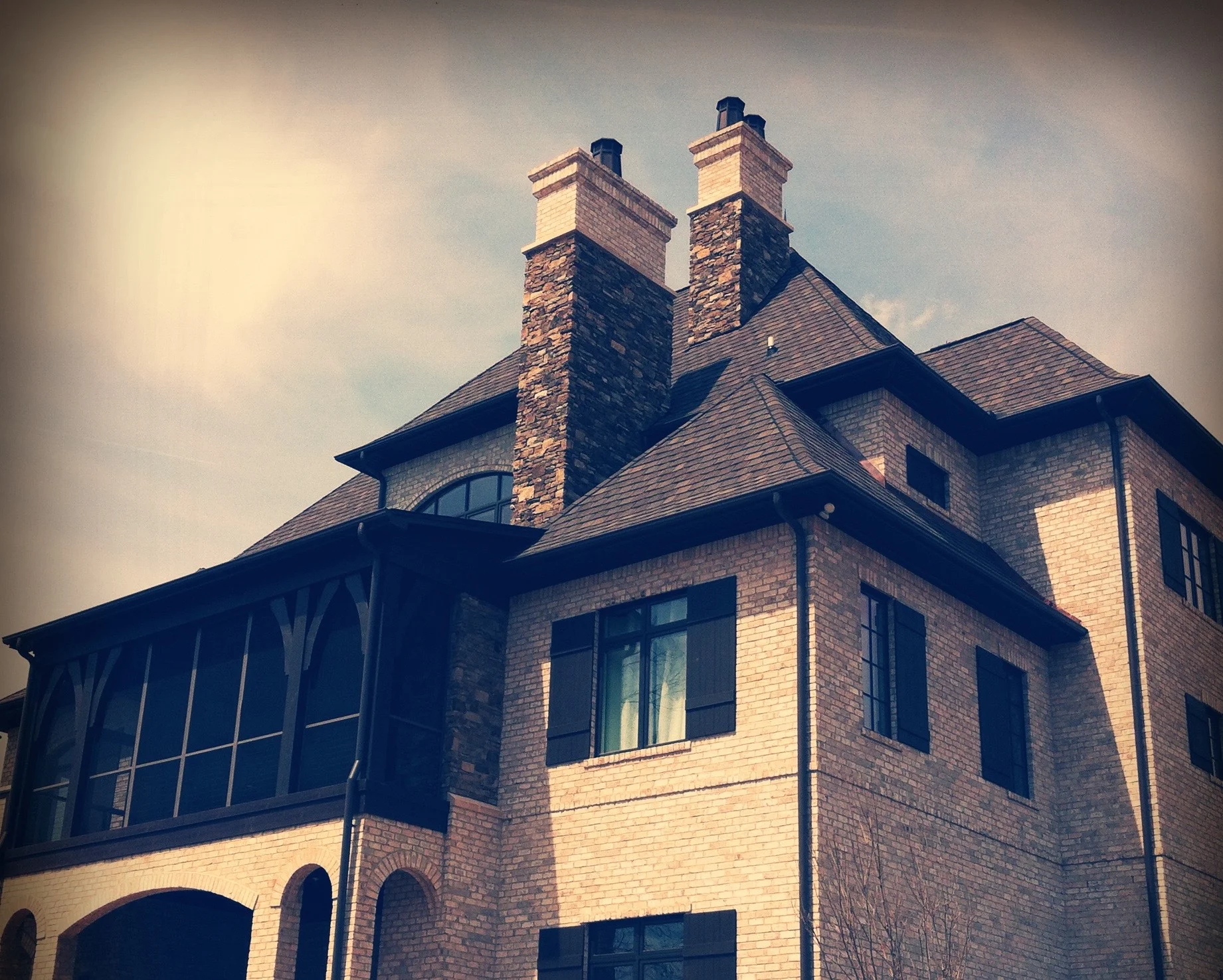 A multi-story brick house with a dark shingled roof, large arched windows, and three prominent brick chimneys against a cloudy sky.