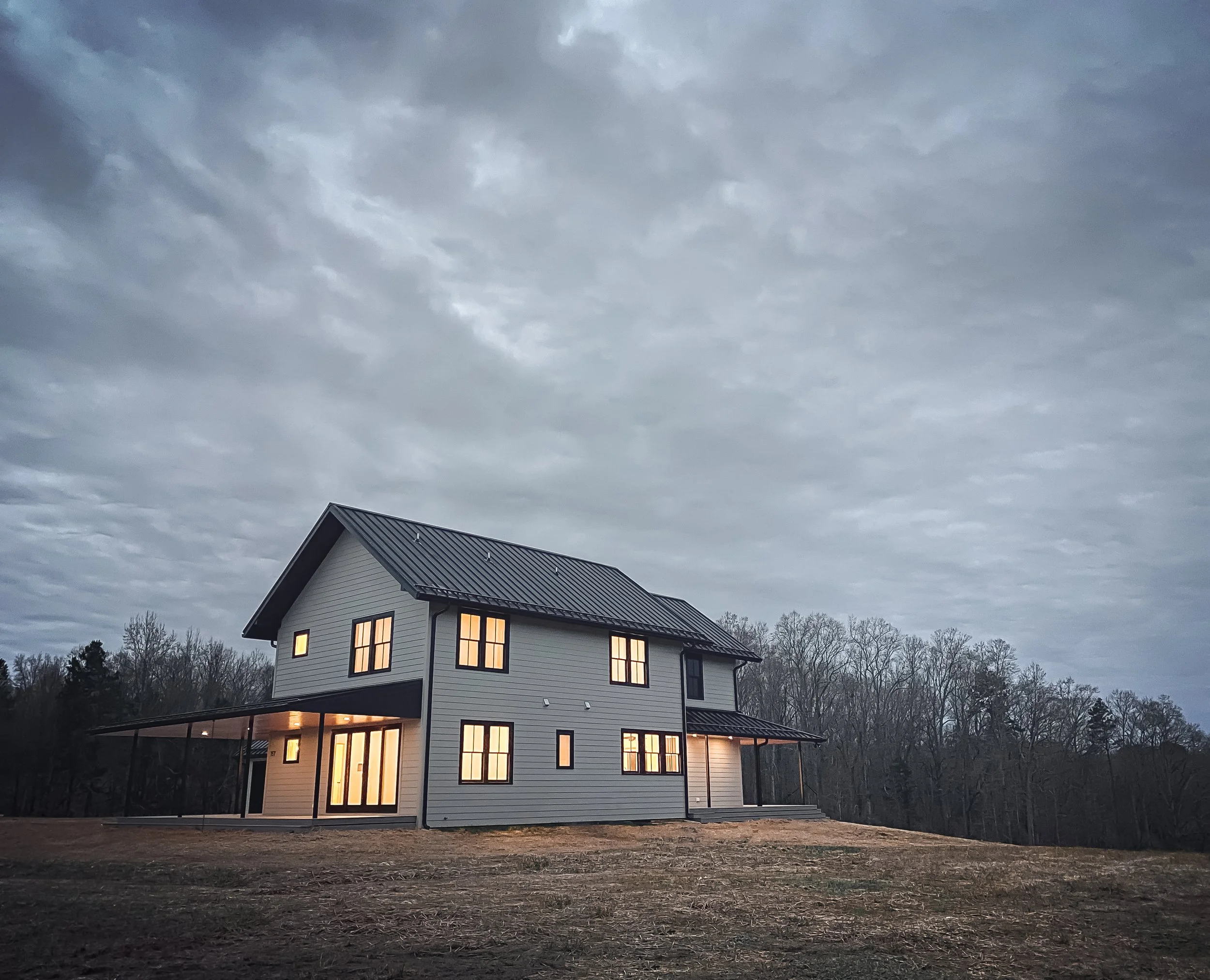 A modern, white, three-story house with large windows and a metal roof, illuminated from inside, set against a cloudy sky with leafless trees in the background.