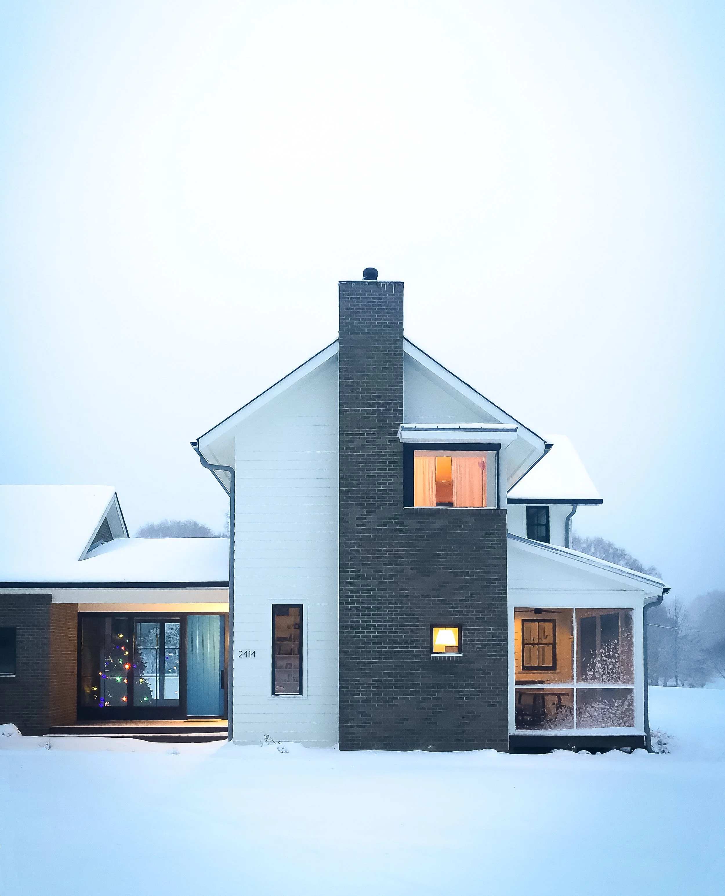 Modern house with snow-covered ground, brick and white siding, and lit windows, one of which shows a decorated Christmas tree inside.