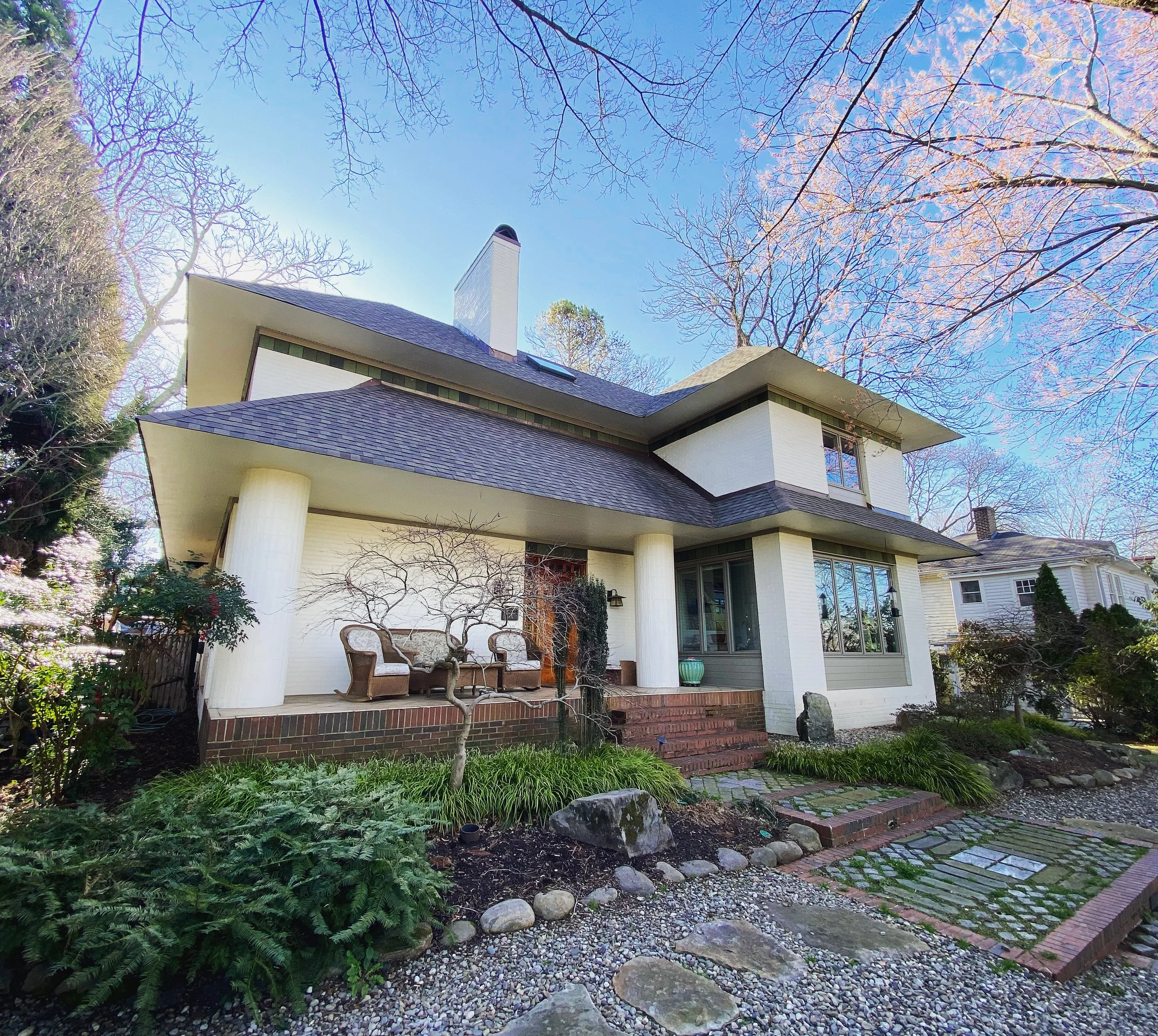 A white house with a sloped roof and a tall chimney, surrounded by leafless trees, shrubs, and a stone pathway leading to the house in a garden setting.