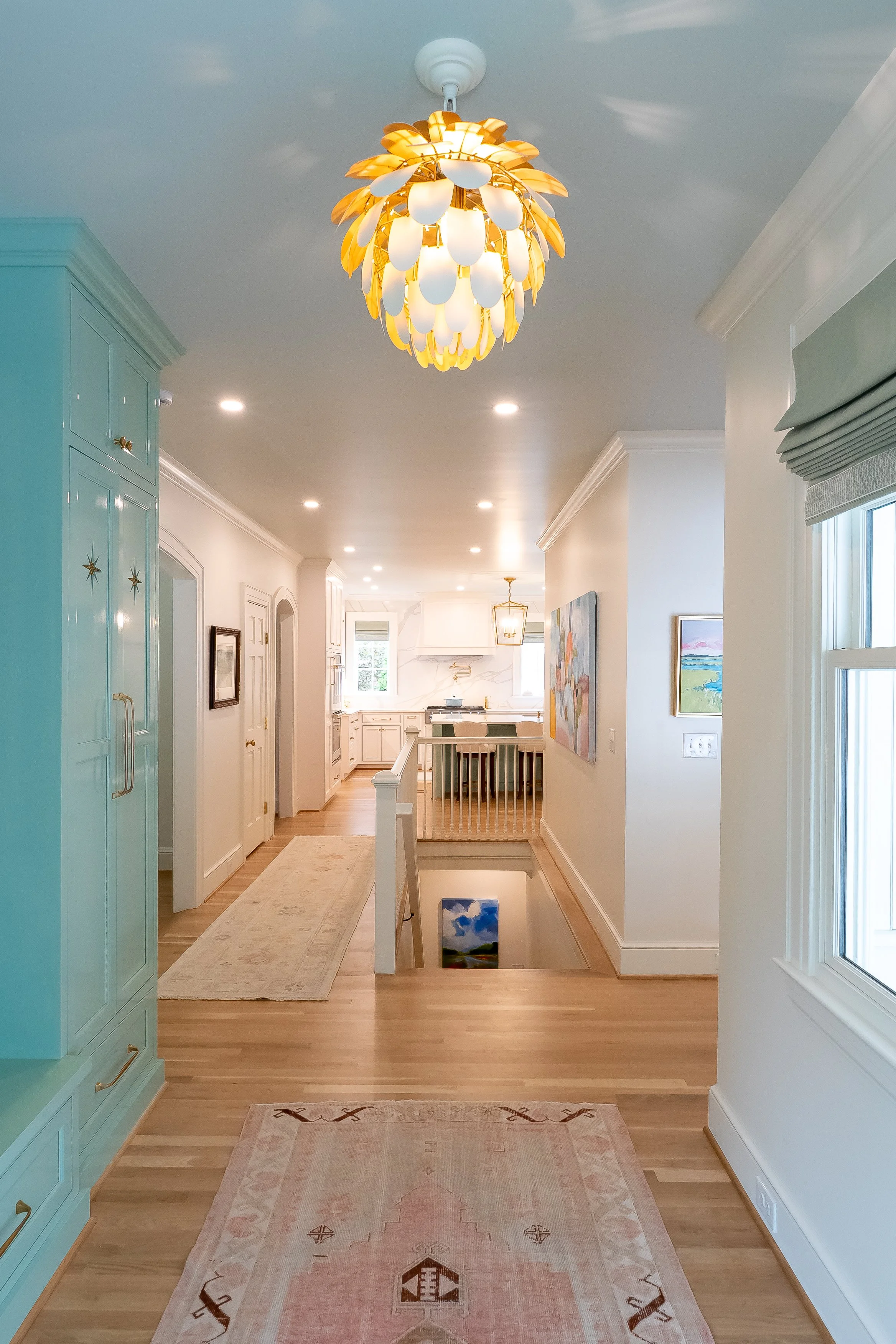 Interior view of a hallway in a house with a modern chandelier, hardwood floors, light-colored walls with framed art, a blue storage cabinet, and a staircase in the distance leading to the lower level.