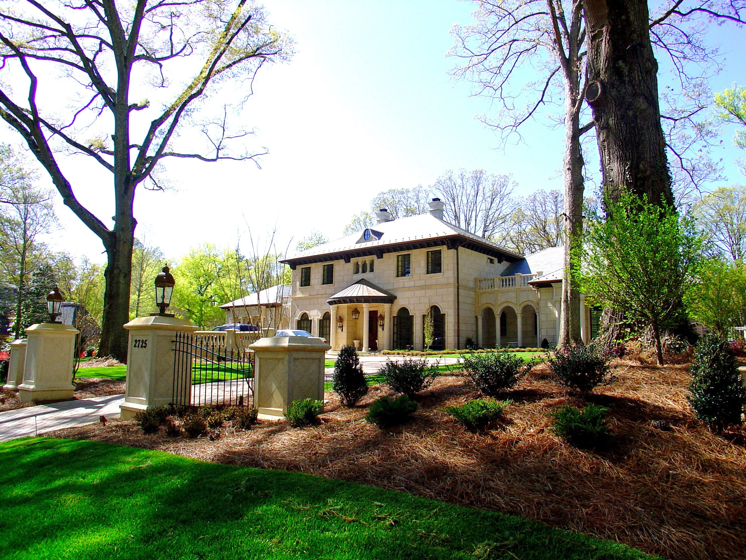 A large house with multiple chimneys and arched windows, surrounded by trees and a garden, with a gate and fence in the foreground.