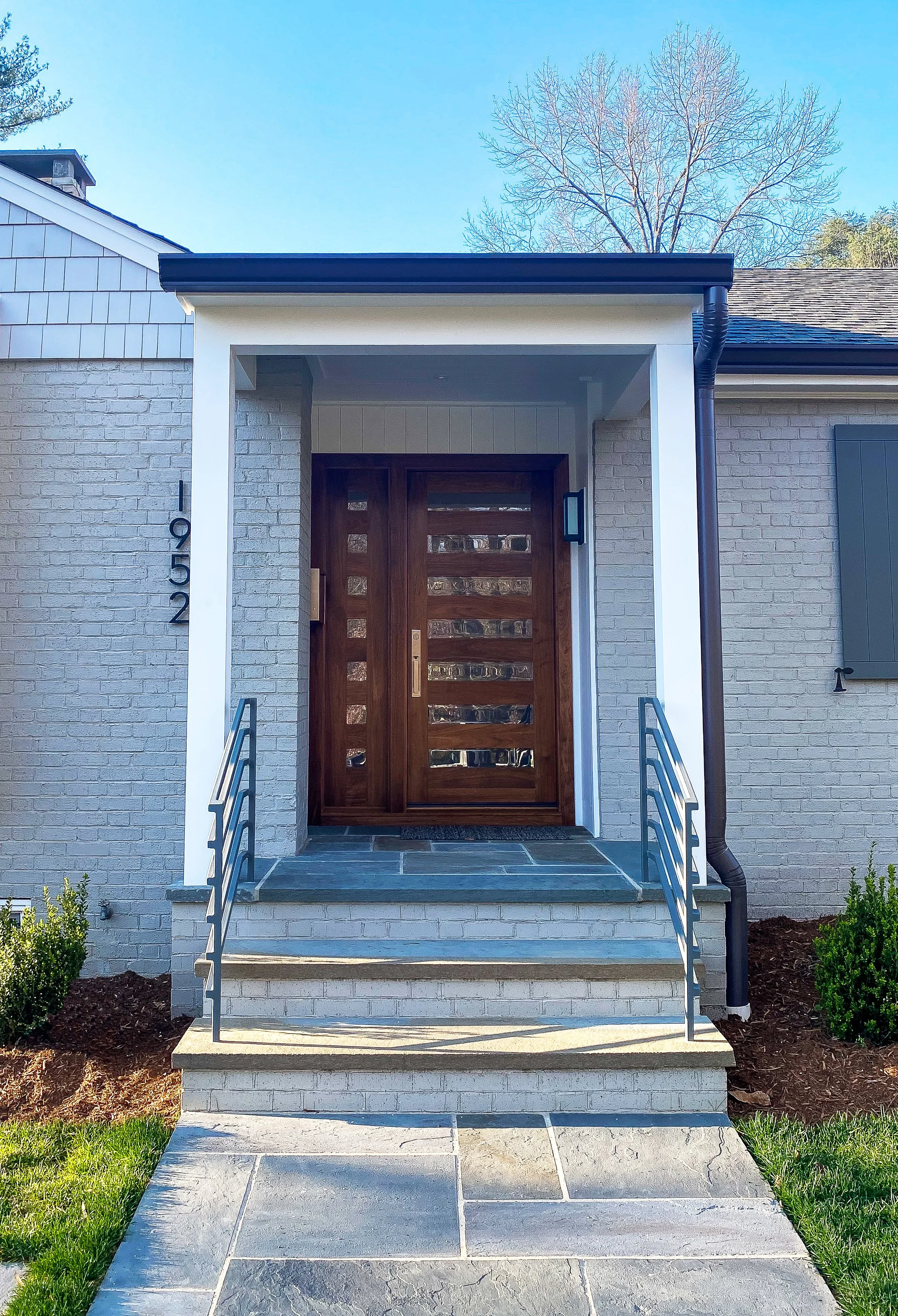Front entrance of a modern house with white brick exterior, wooden door with glass inserts, stone steps, metal handrails, and plants on sides of pathway.