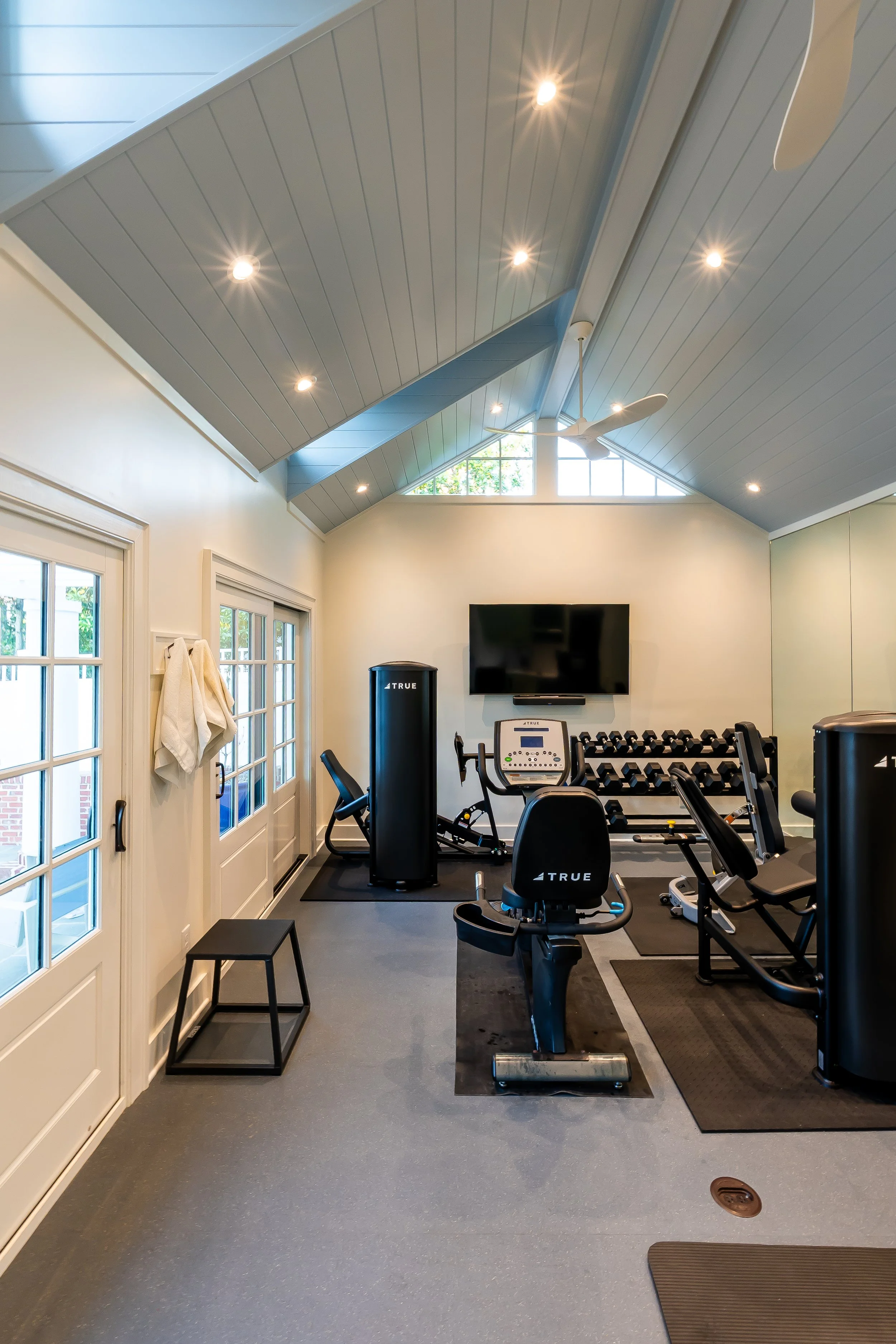 A home gym with exercise equipment including a treadmill, stationary bike, and free weights. There are windows and a door with glass panes, a ceiling fan, and recessed ceiling lights. The room has a gray floor and light-colored walls.