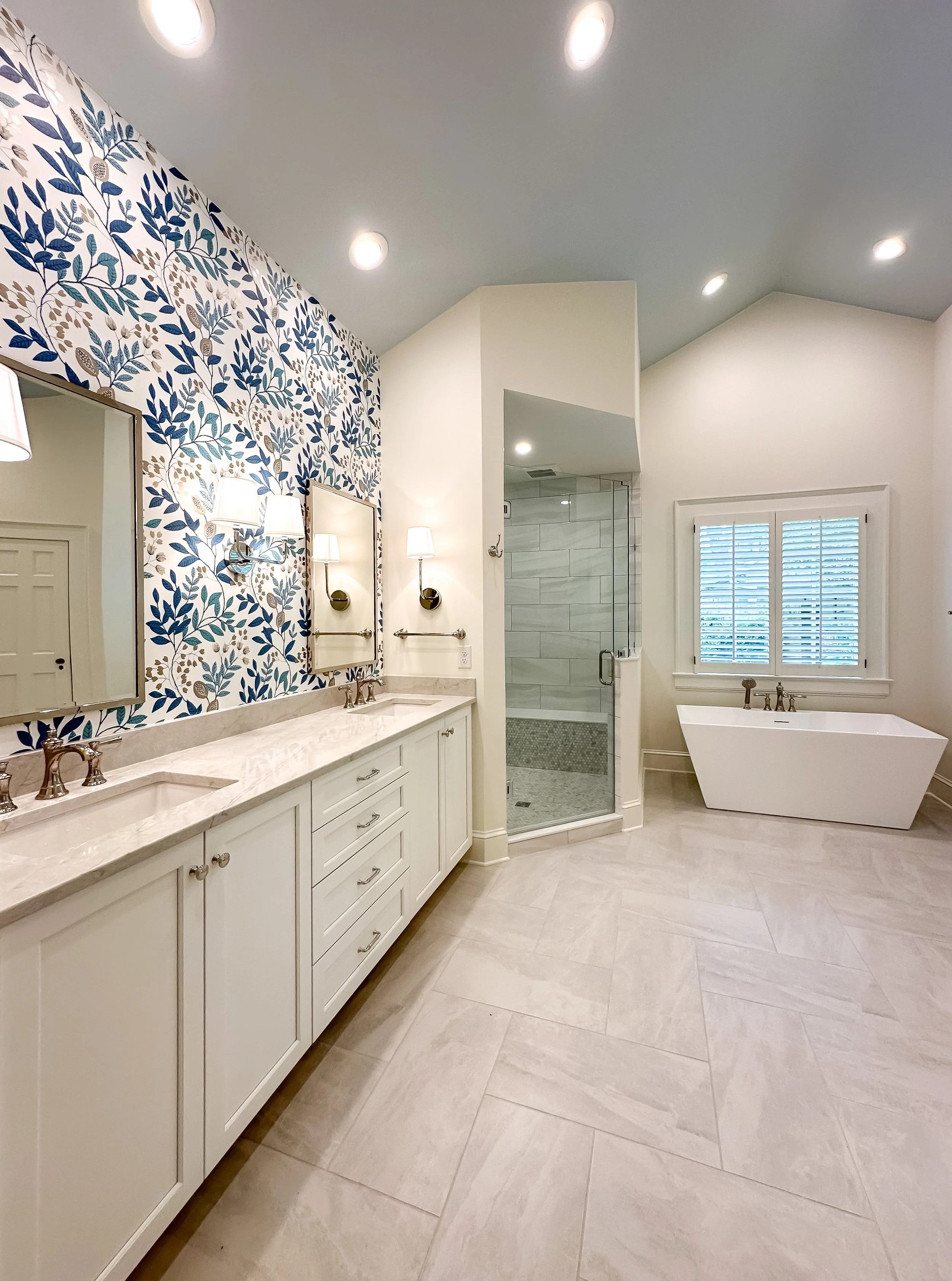 A modern bathroom with a double vanity, framed mirror, wall lamps, a walk-in shower with glass door, a window with shutters, and a freestanding bathtub.