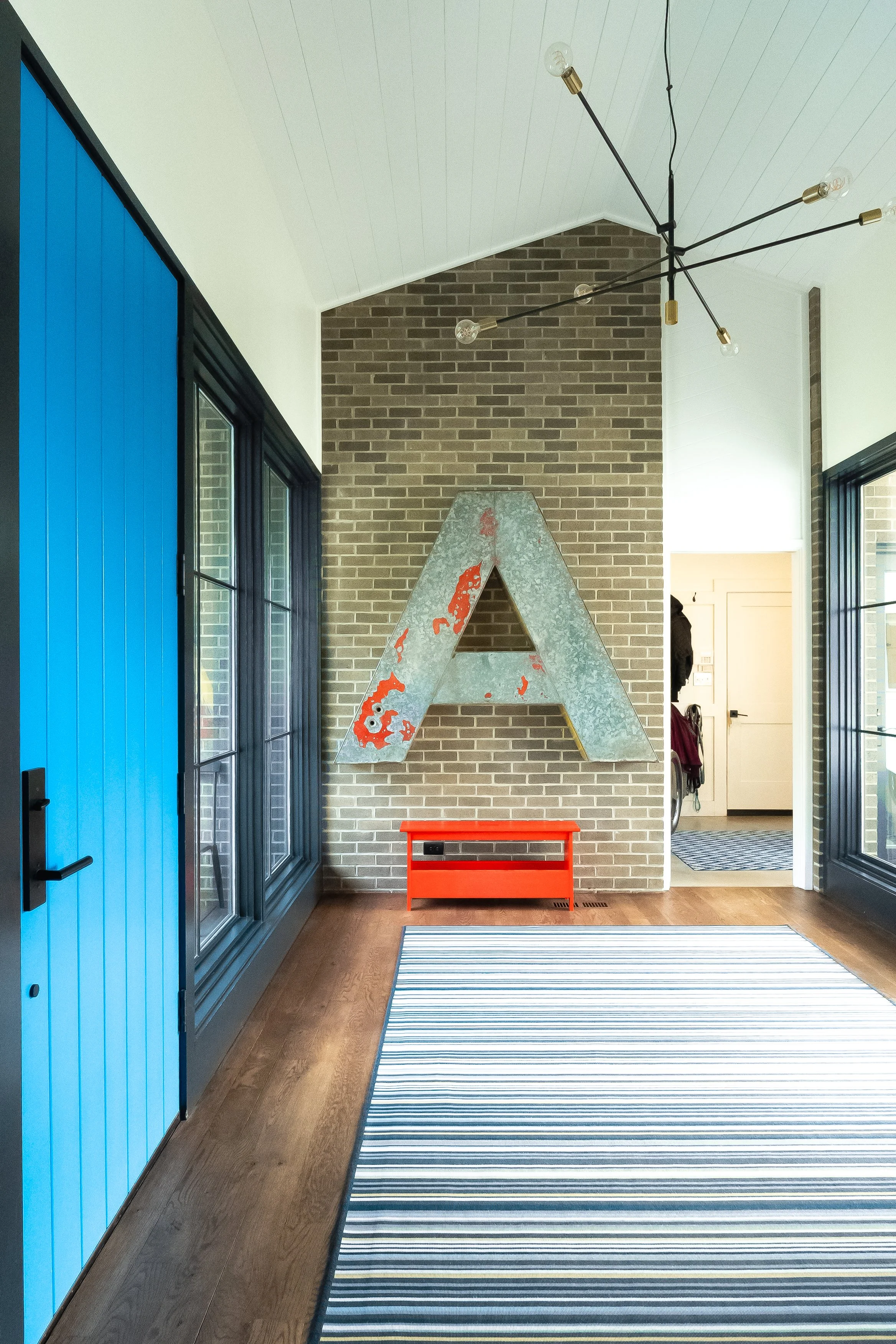 Entryway with blue door on left, large windows, striped rug, a large metal letter A on the brick wall, a small red table underneath, and a view to the hallway and other rooms.