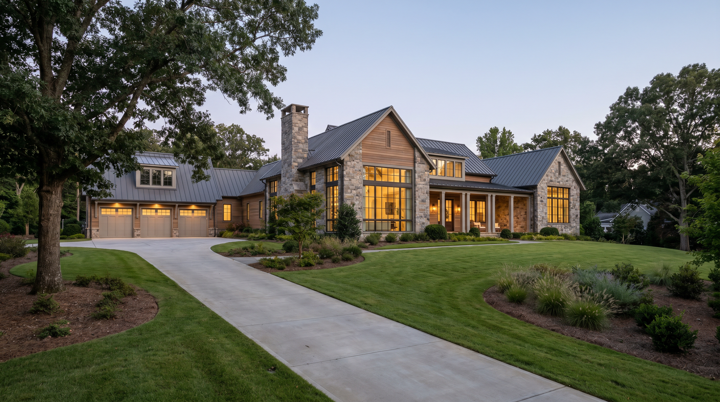 Modern house with large glass windows, stone and wood exterior, and metal roof, surrounded by green lawn and trees, at dusk.