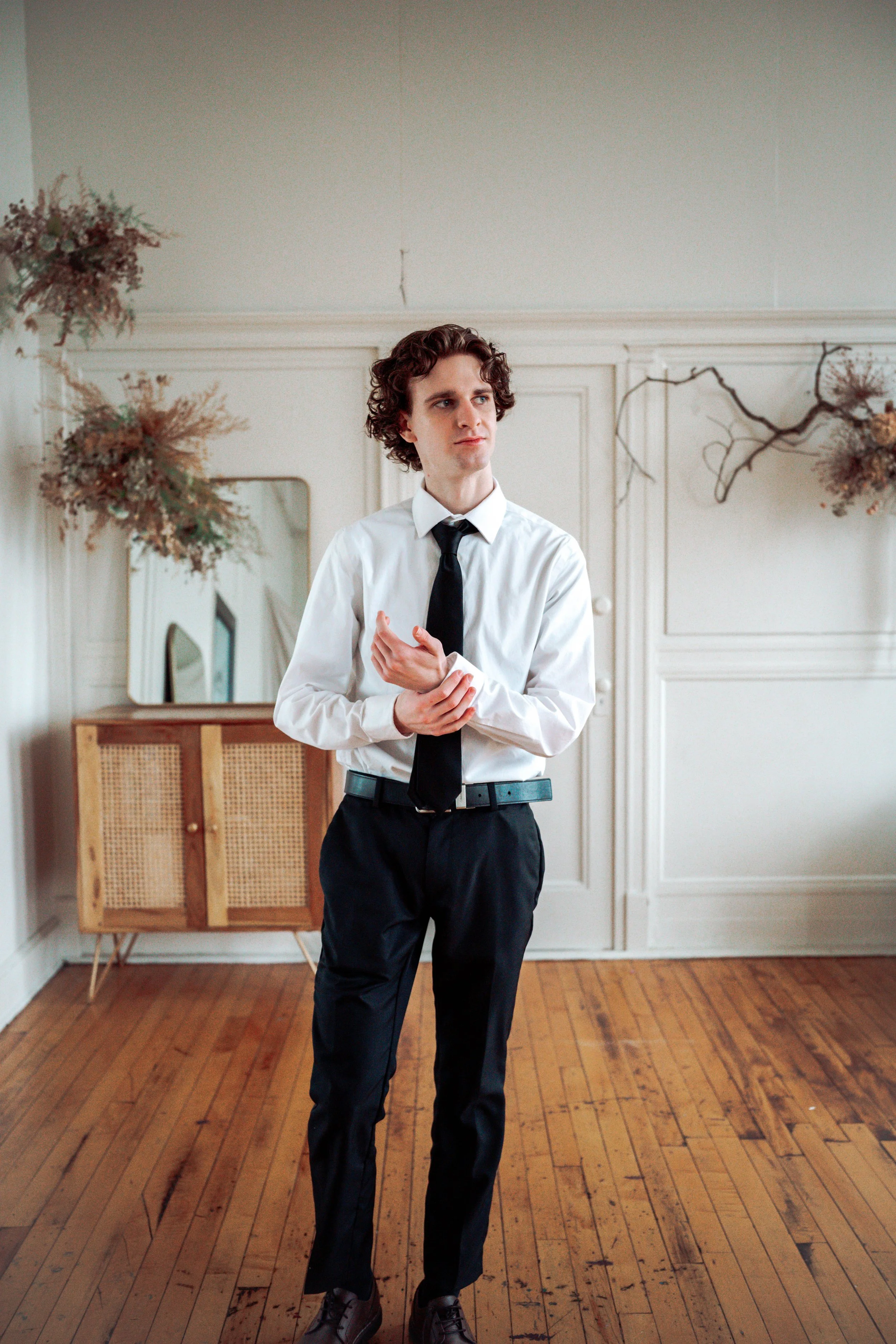 A young man in a white dress shirt, black tie, and black pants standing in a room with wooden flooring and white walls. He is adjusting his cufflinks, looking to the side with a thoughtful expression.