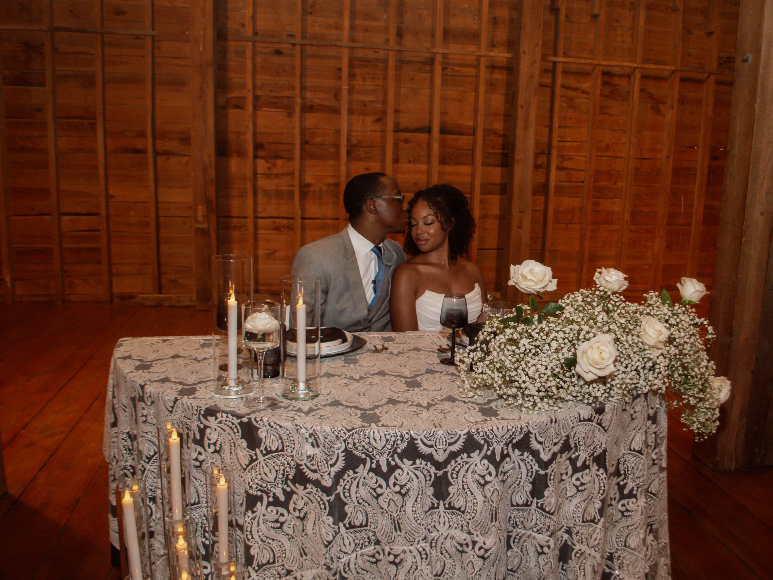 A couple sitting at a wedding reception table with candles, wine glasses, and white roses, in a wooden venue, sharing a romantic moment.
