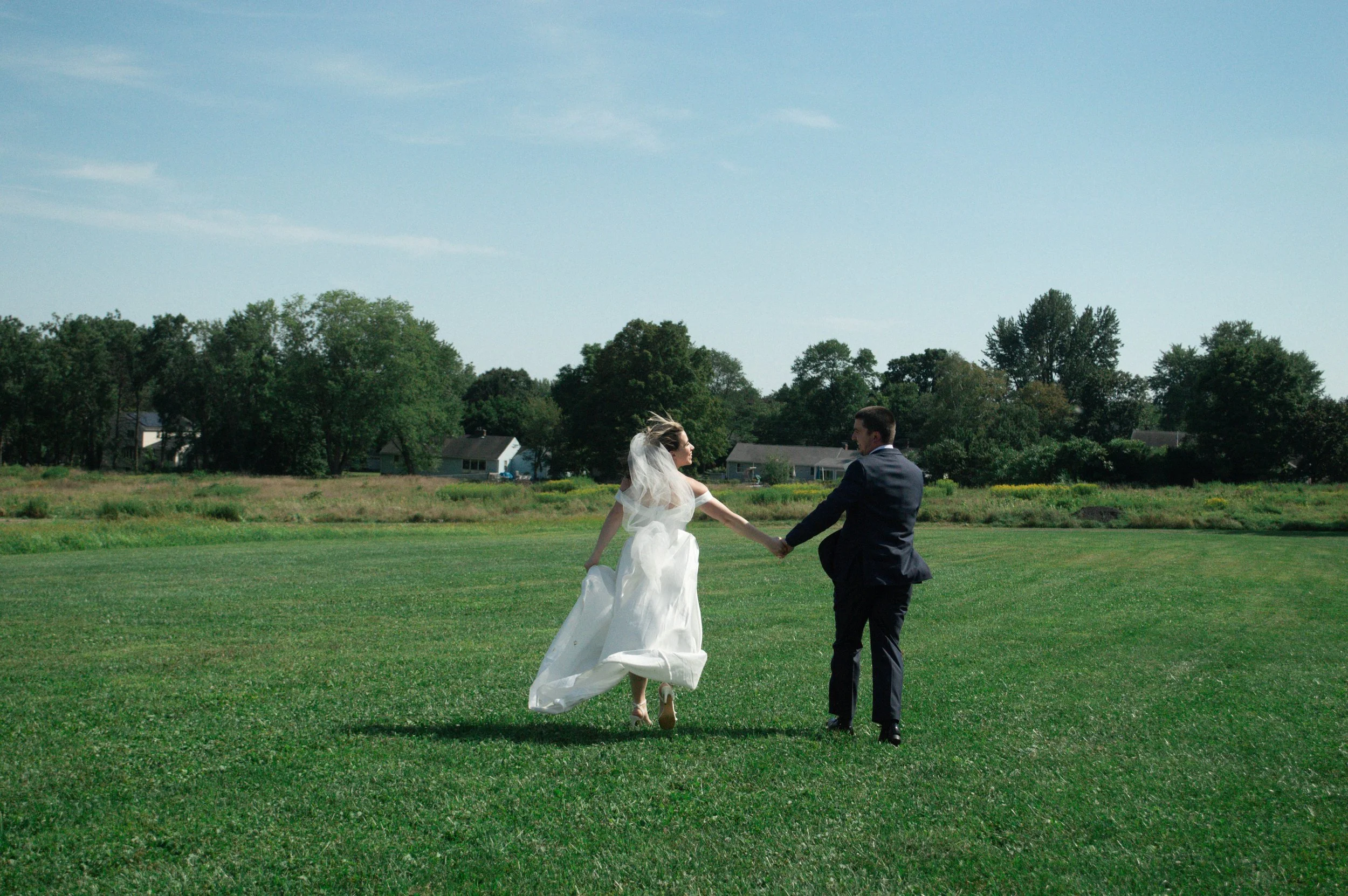 A bride and groom are holding hands and walking across a grassy field on a sunny day.