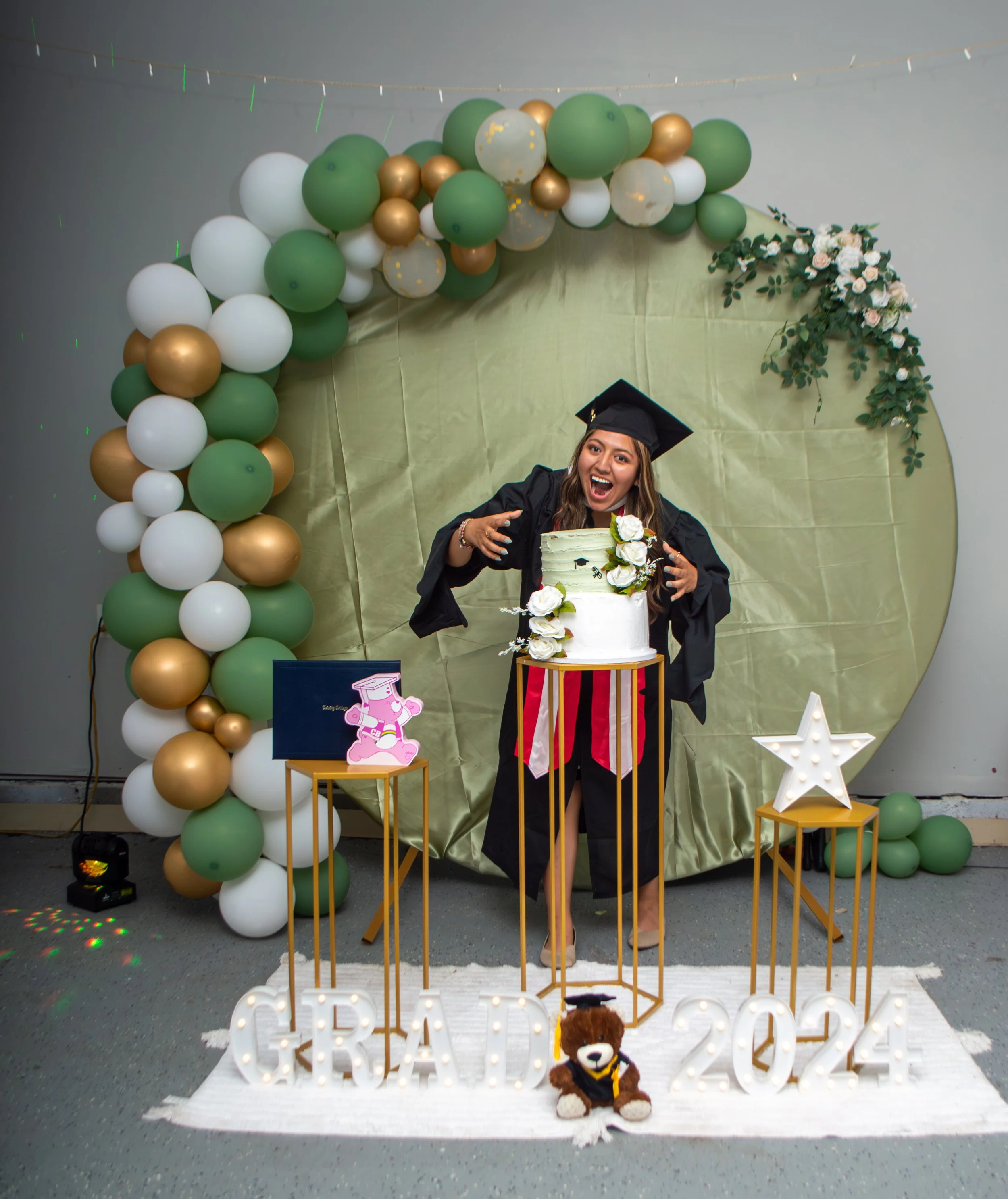 A woman in a graduation cap and gown celebrating her graduation with a cake, surrounded by balloons, decorations, and a sign that reads 'GRAD 2024'.