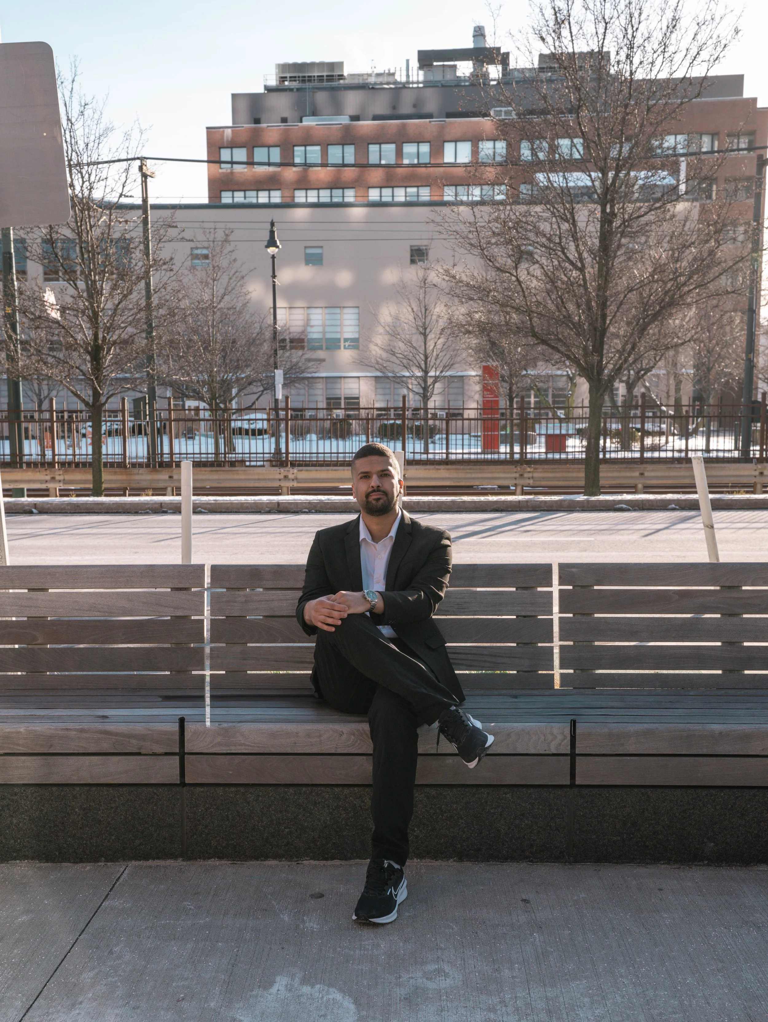 A man in a black suit sitting on a wooden bench outdoors with city buildings, trees, and a street in the background