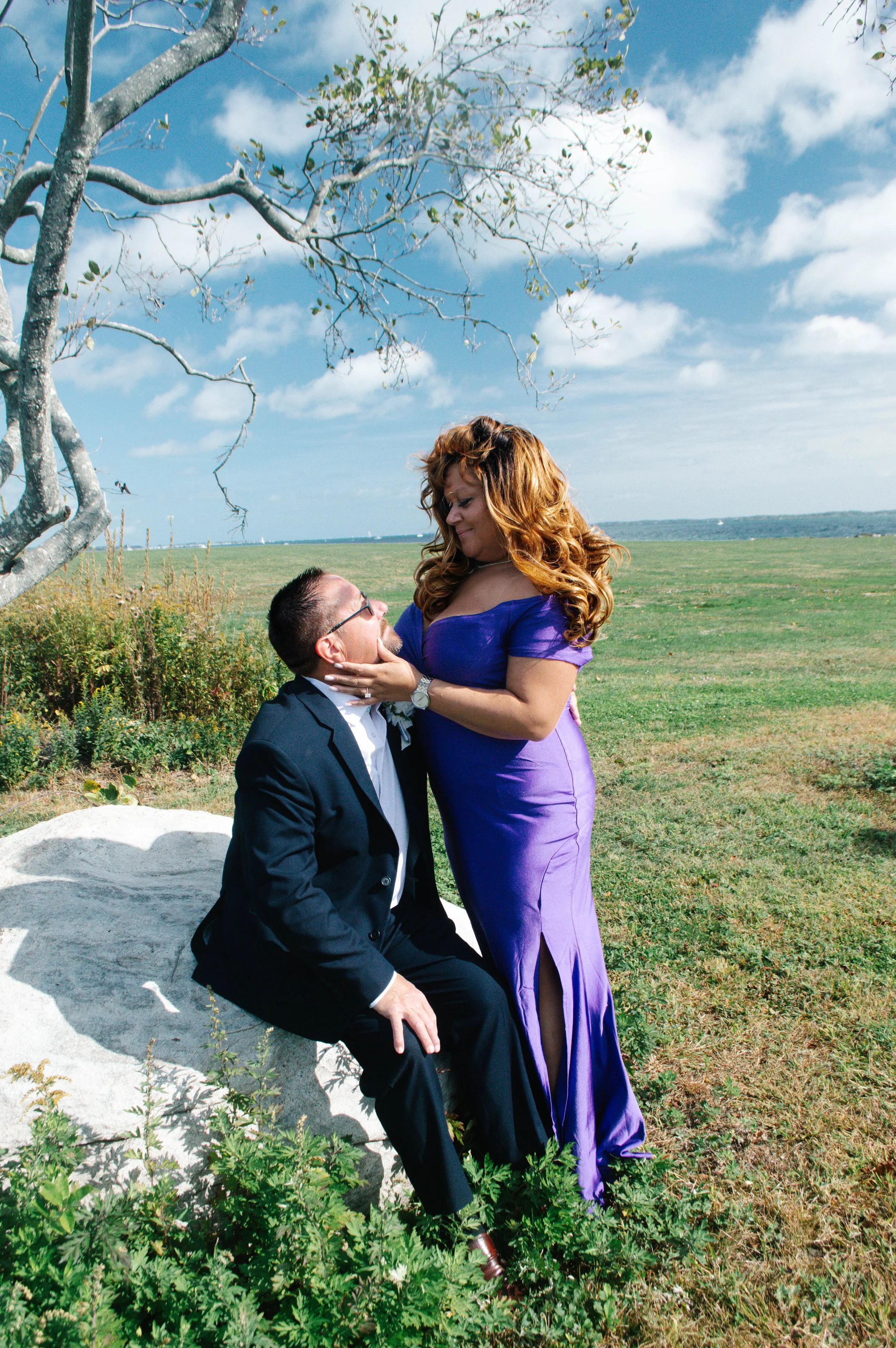 A man in a suit and a woman in a purple dress share an intimate moment outdoors by a tree on a sunny day with green grass and a blue sky with clouds.