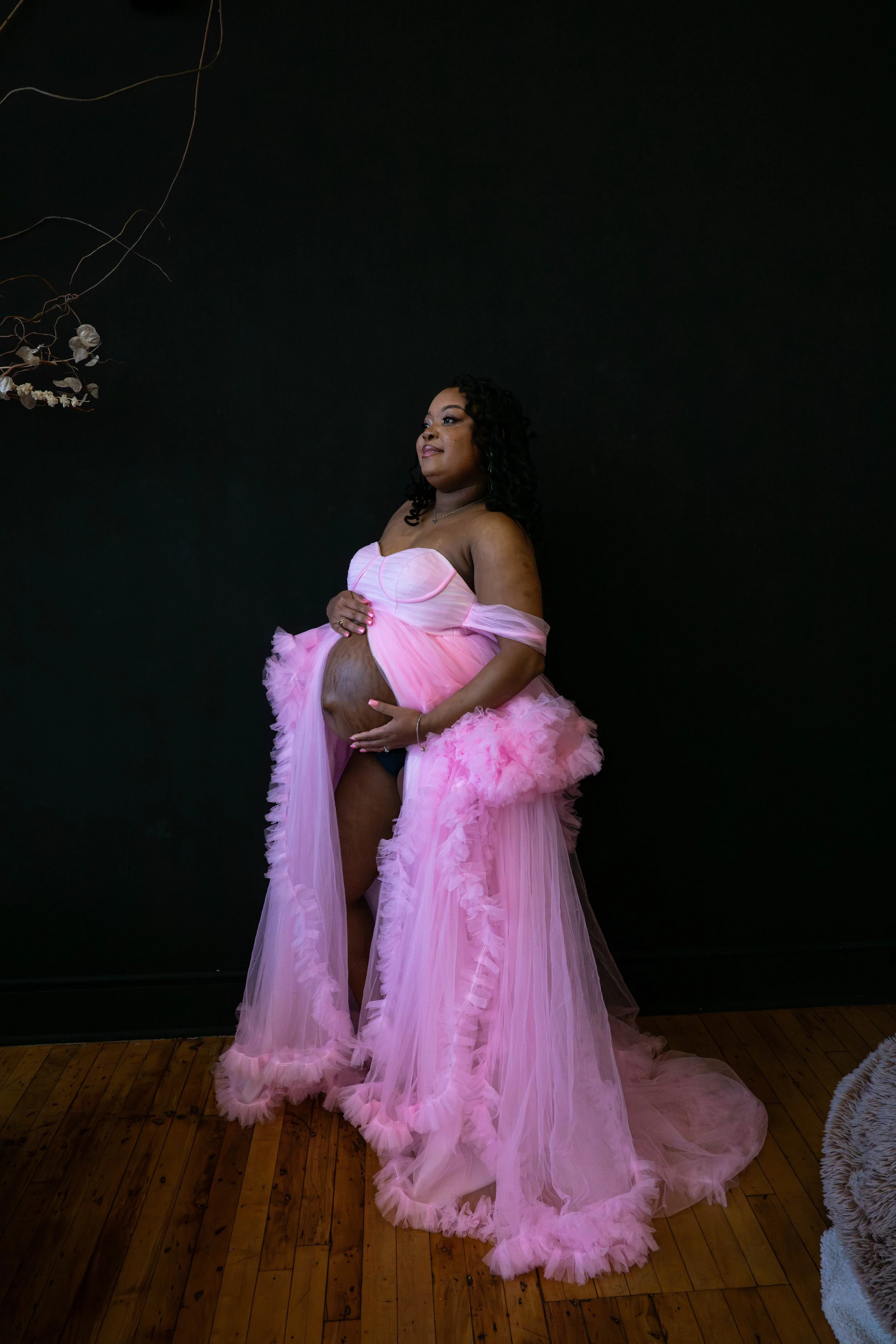 Pregnant woman in a pink, tulle gown standing against a dark wall, holding her belly and looking upward.
