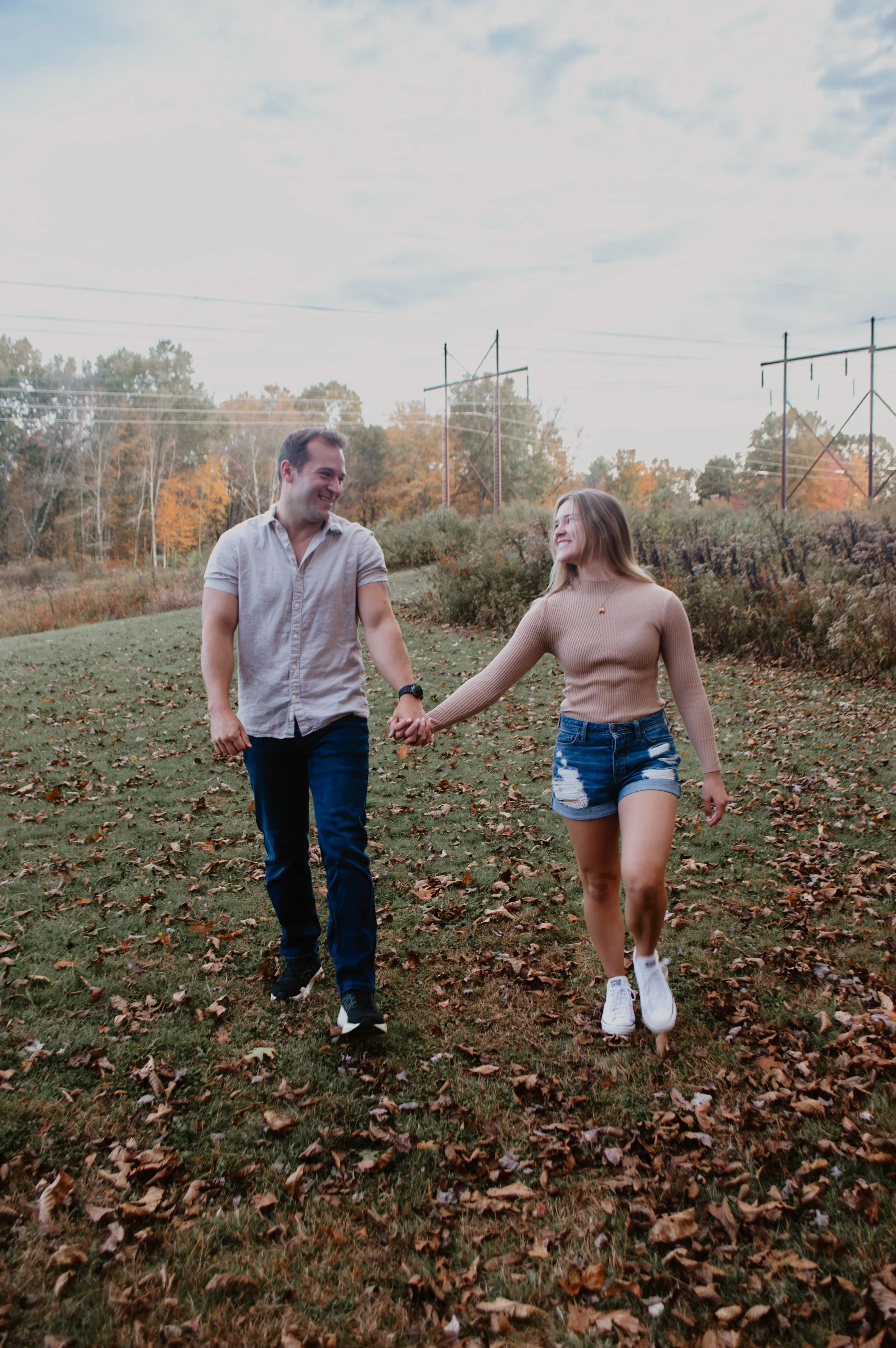 A young couple walking hand in hand through a park with fallen leaves, trees in autumn colors, and power lines in the background.