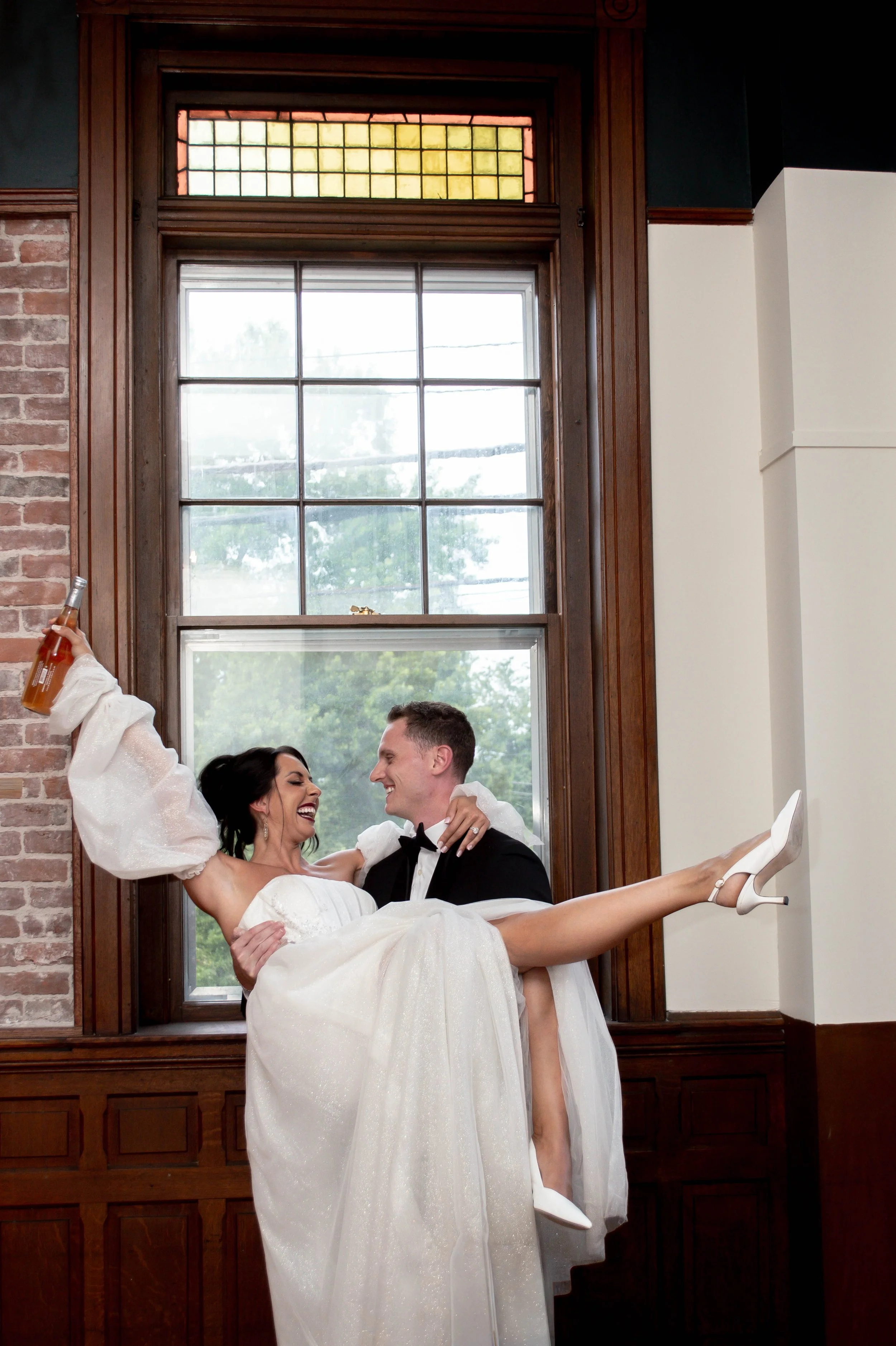 A joyful bride being carried by her groom in a dimly lit room with large stained glass windows and brick wall detail, she holds a beer bottle and wears a white wedding gown and heels.
