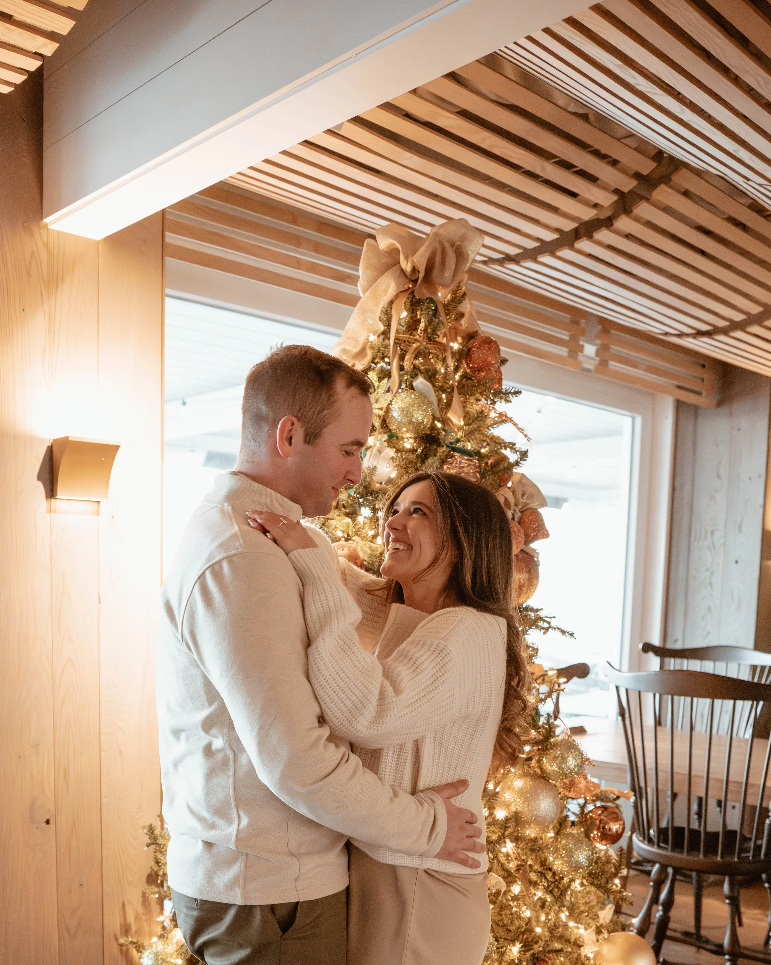 A couple embraces in front of a decorated Christmas tree, with a wooden wall and large window in the background.