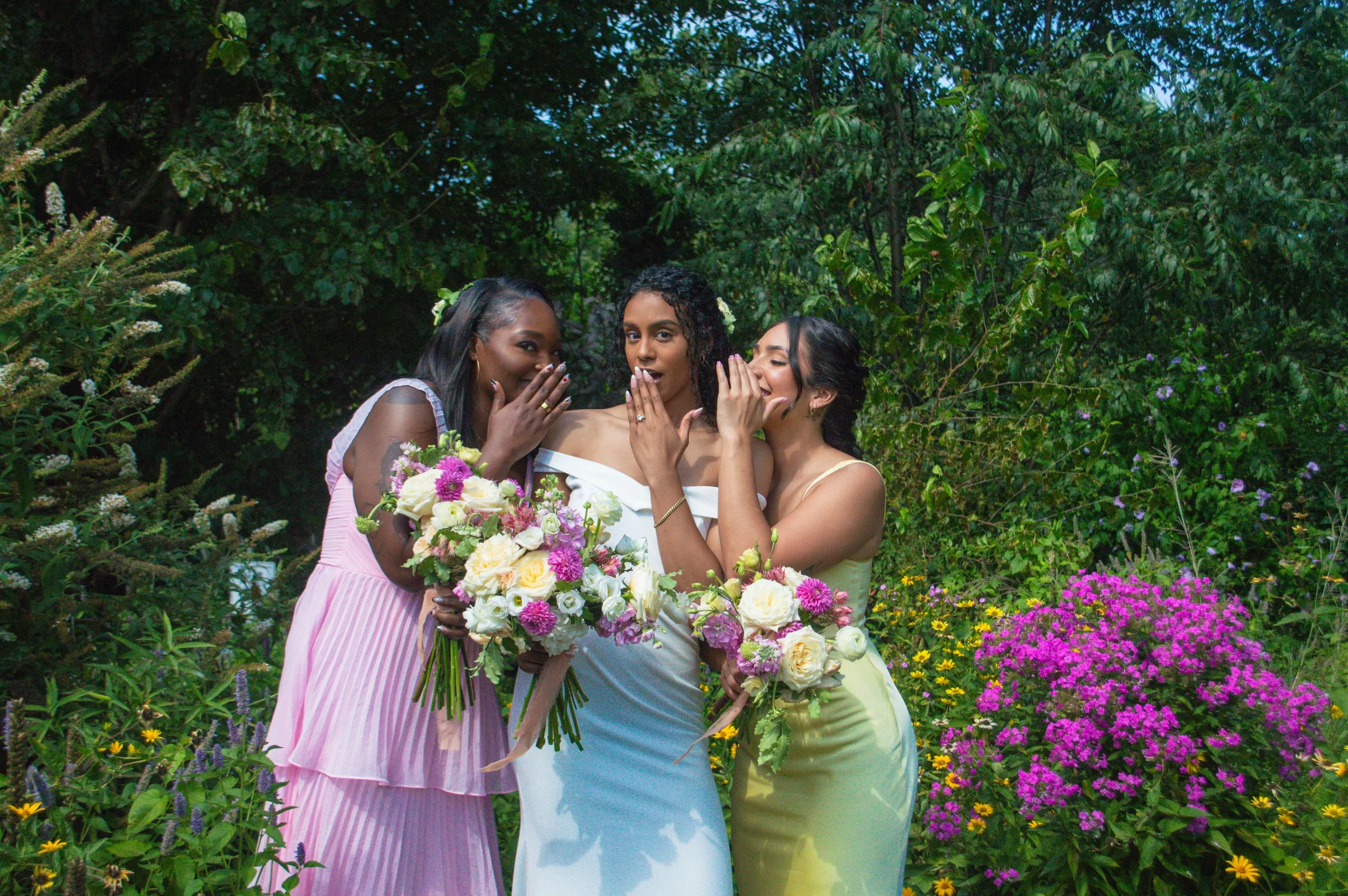 Three women in colorful dresses standing together in a lush garden with vibrant flowers, each holding a bouquet of flowers, celebrating a special occasion.