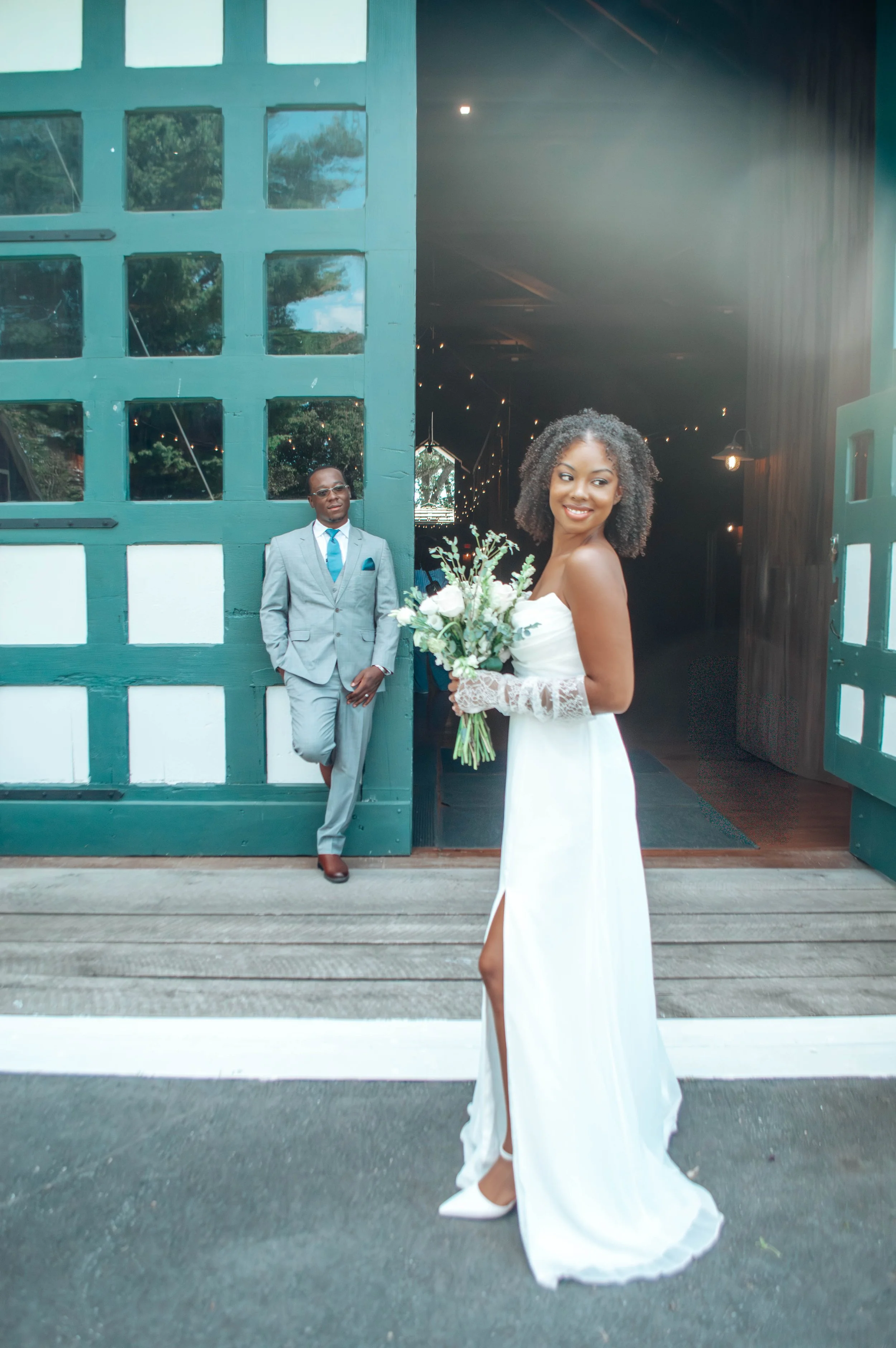 A bride holding a bouquet of white flowers with lace gloves, standing outside near a green barn-style door, while a groom in a gray suit and blue tie leans against the doorframe in the background.