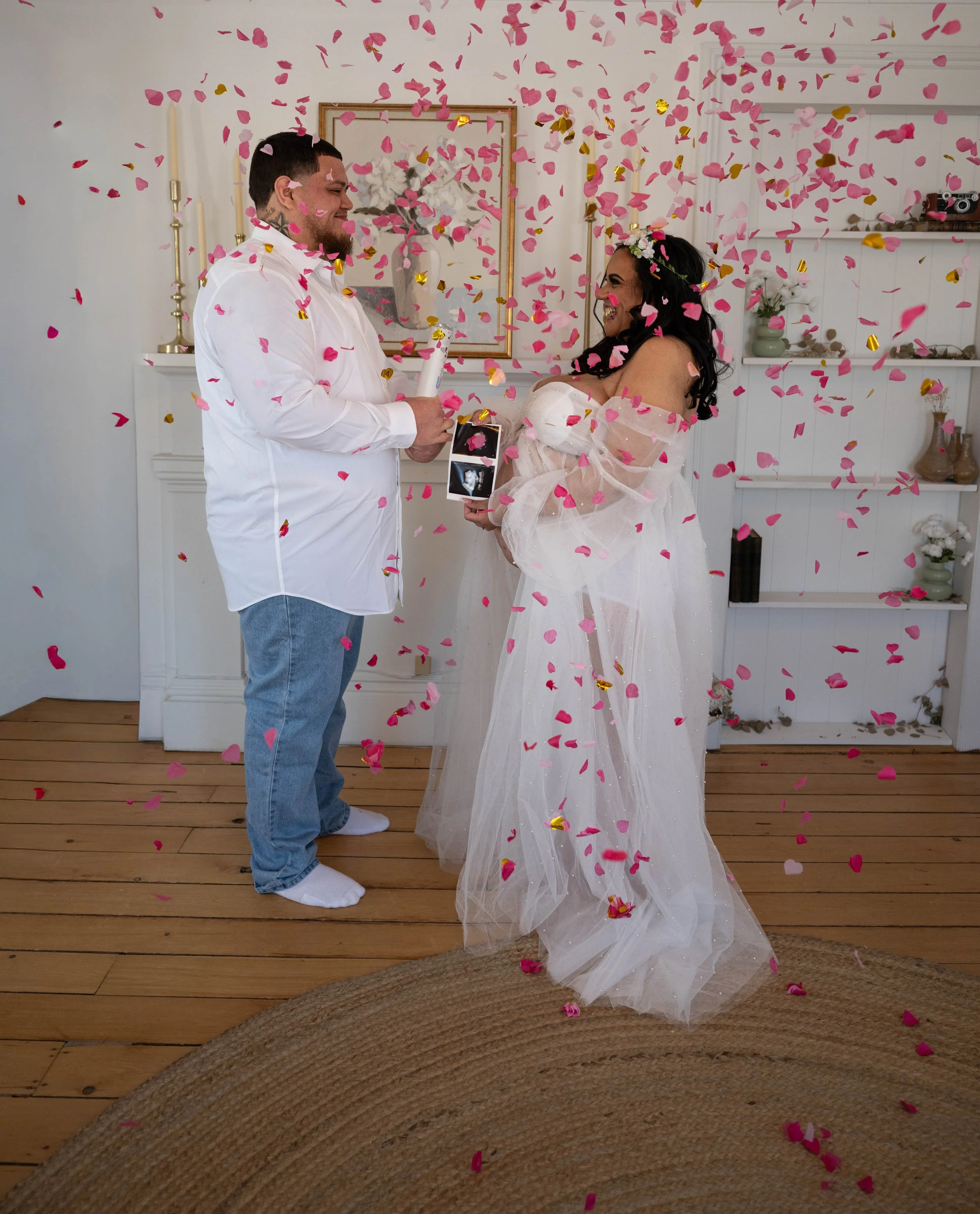A couple stands in a room with wooden floors and white walls, celebrating with pink and gold confetti falling around them. The man is wearing a white shirt and jeans, holding a small cake, while the woman is dressed in a sheer white gown with a flowe