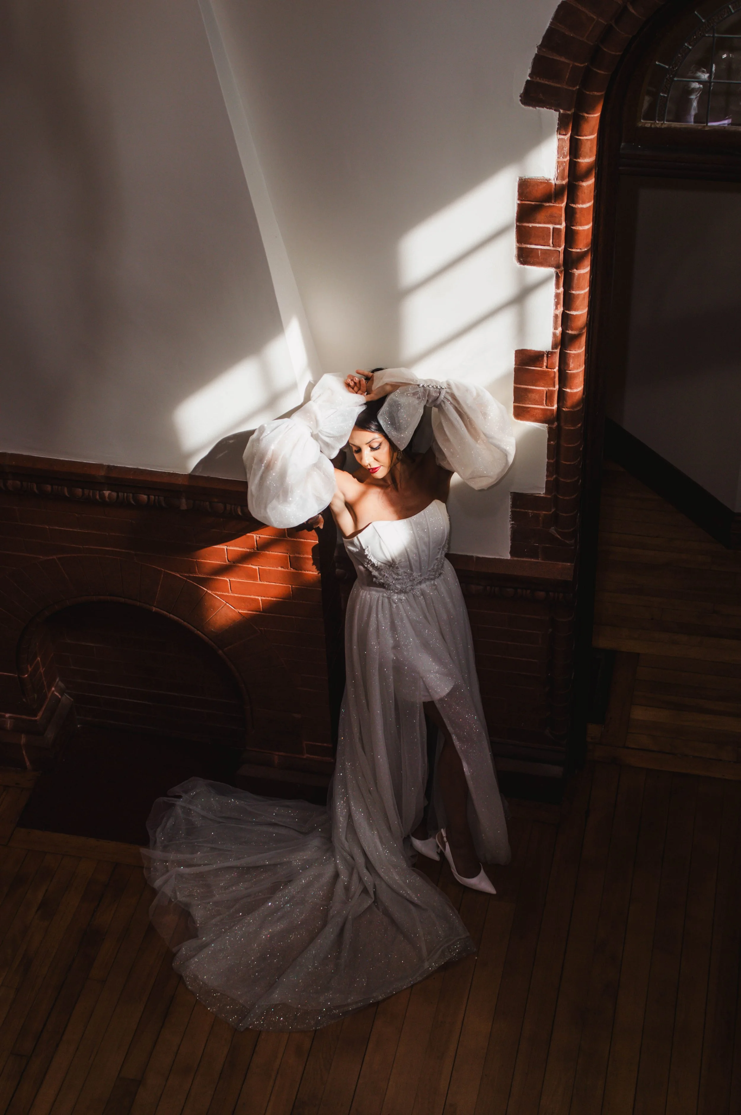 A woman in a glittery silver dress and high heels stands beside a brick fireplace, basking in sunlight streaming through a nearby window.