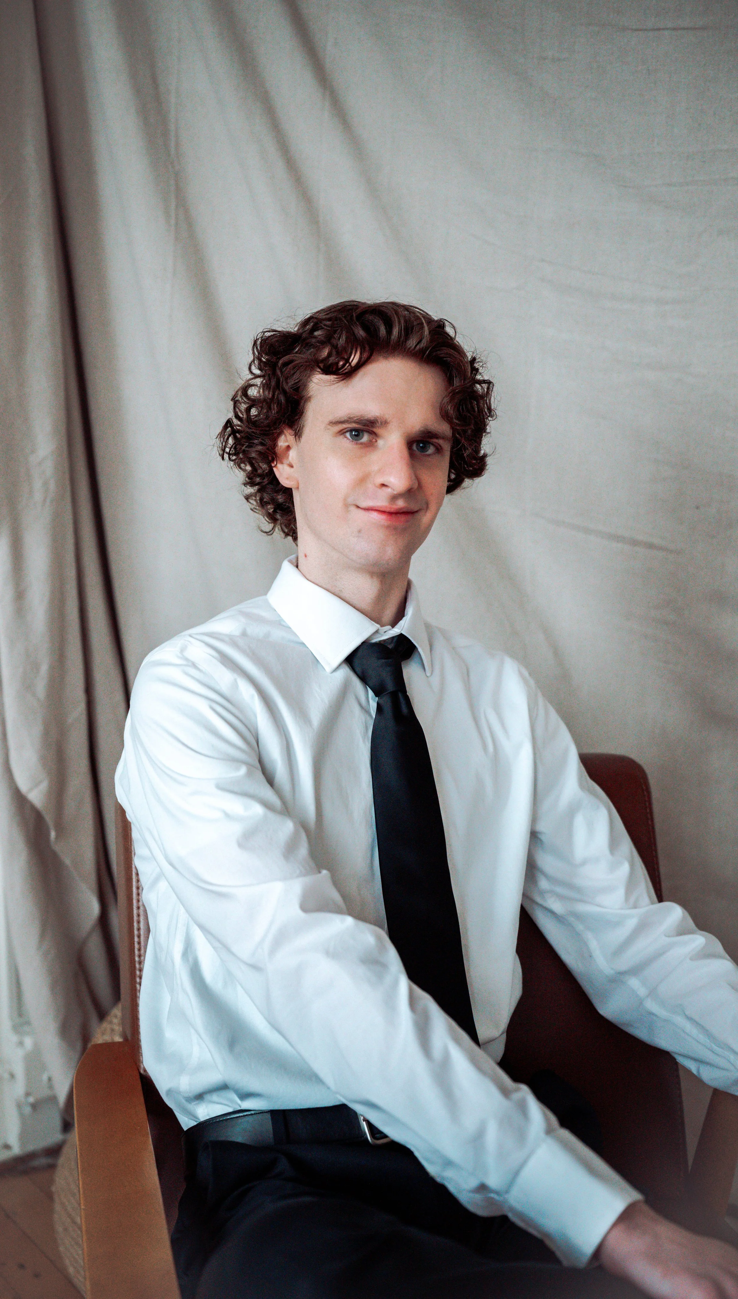Young man with curly brown hair and blue eyes, dressed in a white shirt and black tie, sitting on a chair with a neutral background.