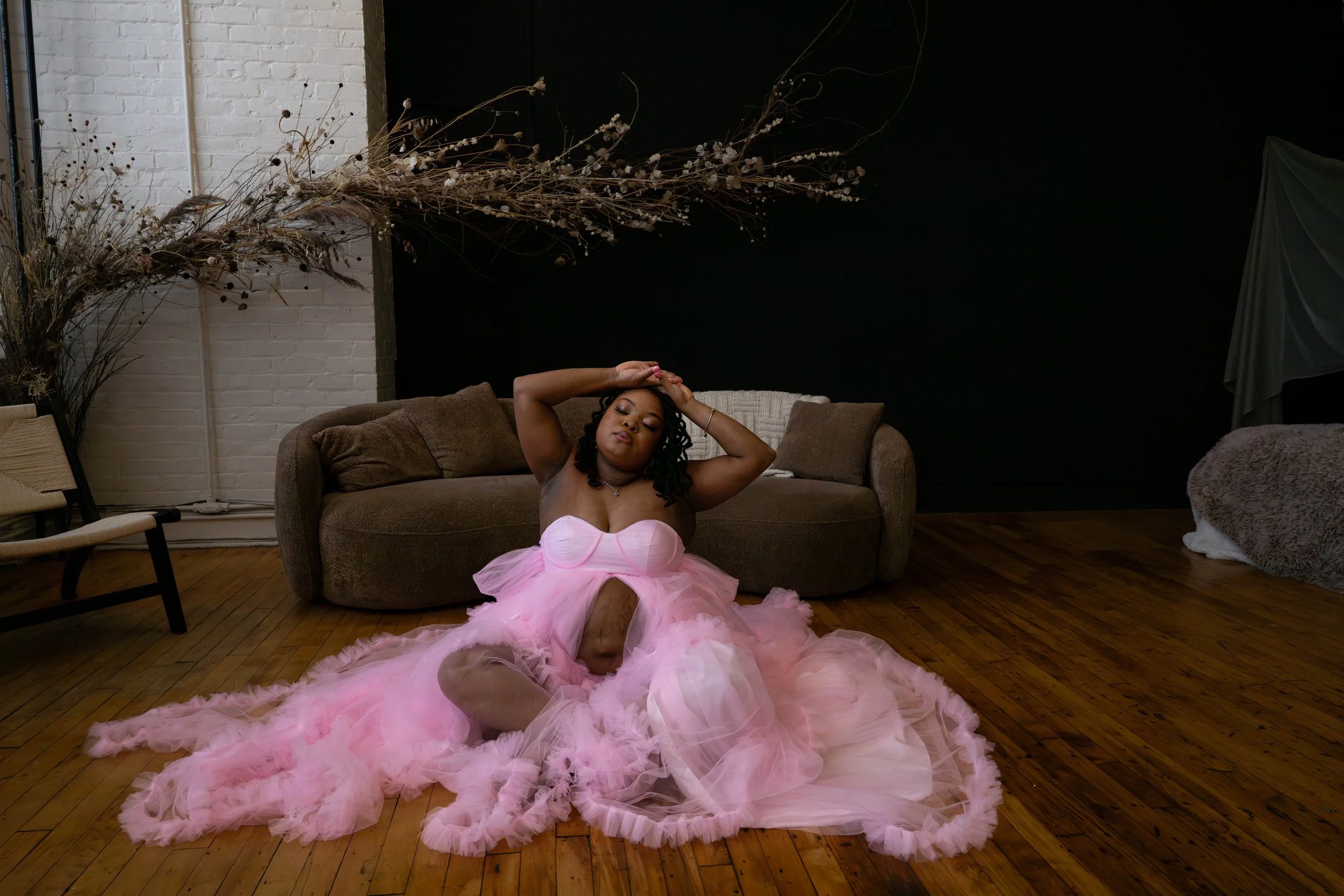 A woman sitting on the hardwood floor wearing a pink tulle dress with a strapless bodice and a full skirt, striking a pose with her eyes closed and hands resting on her head, in a room with a brown sofa, white brick wall, and dark wall background.
