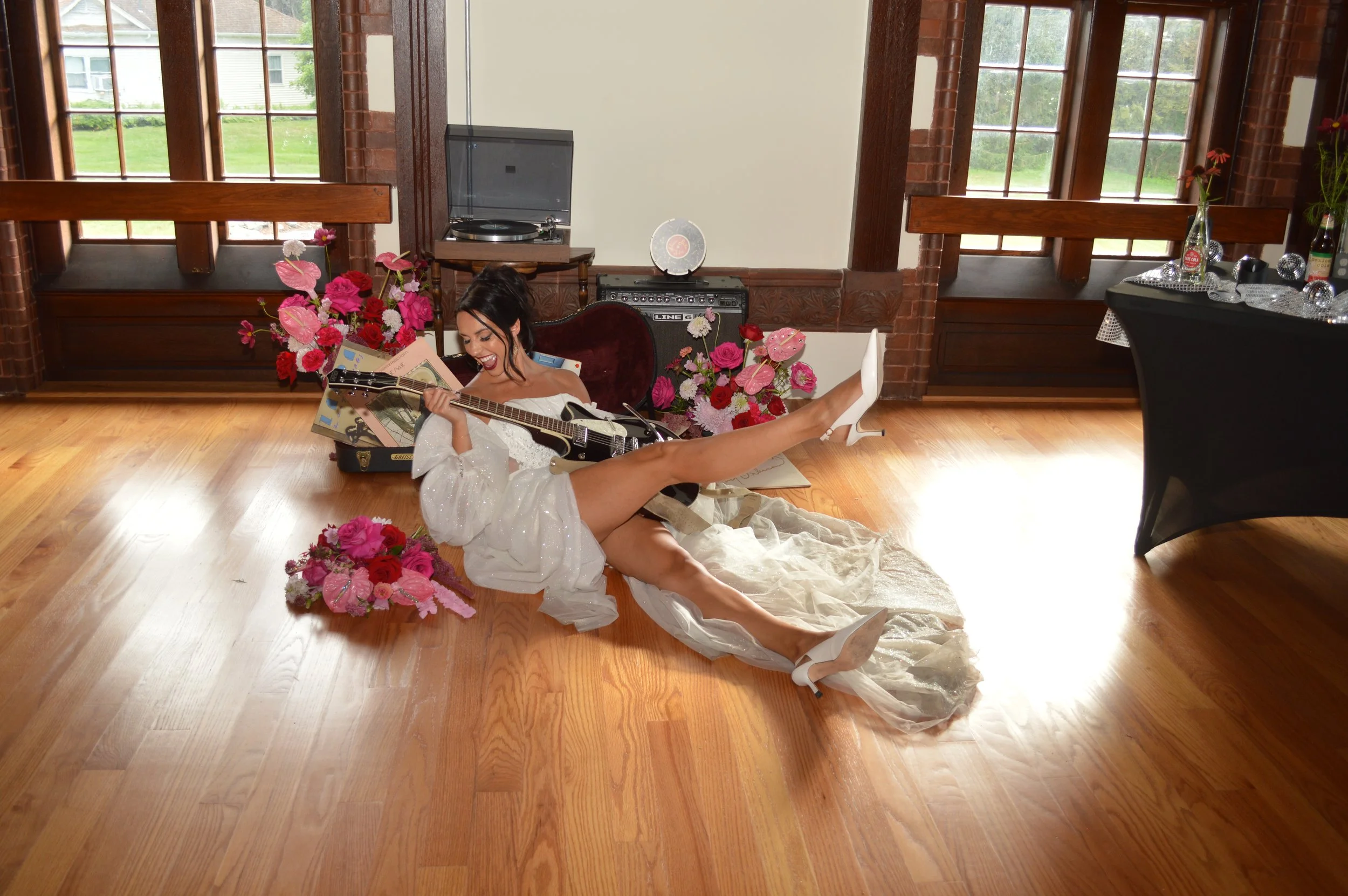 A woman in a white dress and high heels playing guitar on wooden floor surrounded by colorful flowers and decorations in a bright room with large windows.