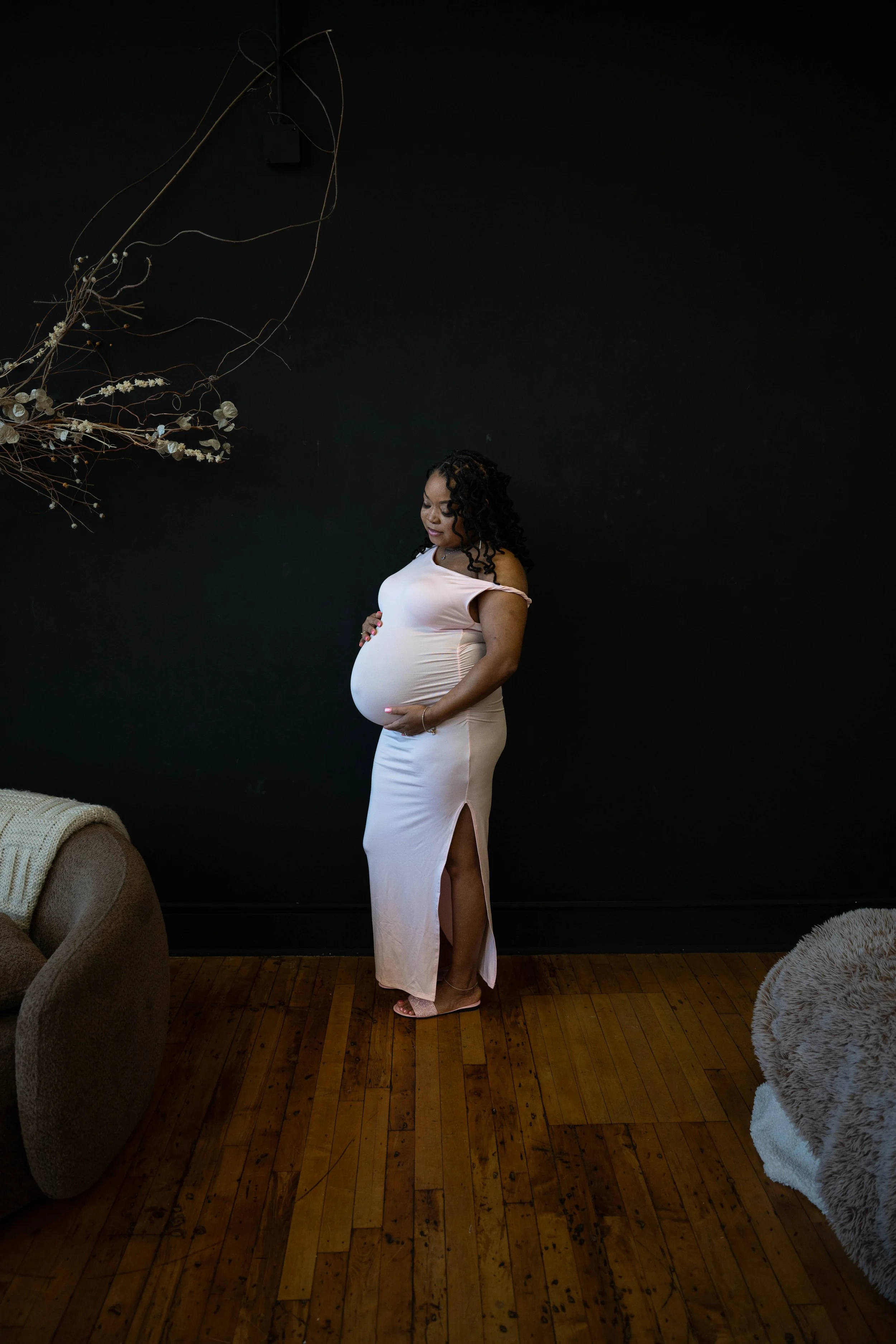 A pregnant woman standing against a black wall, wearing a light pink dress with a slit on the side, looking down at her belly, with curly hair and surrounded by minimalistic decor.