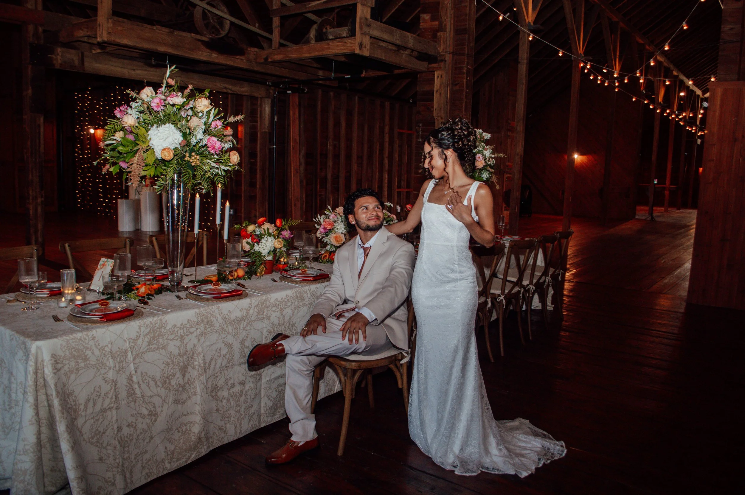 A bride and groom at their wedding reception with decorated table and floral centerpieces, inside a rustic wooden venue with string lights.