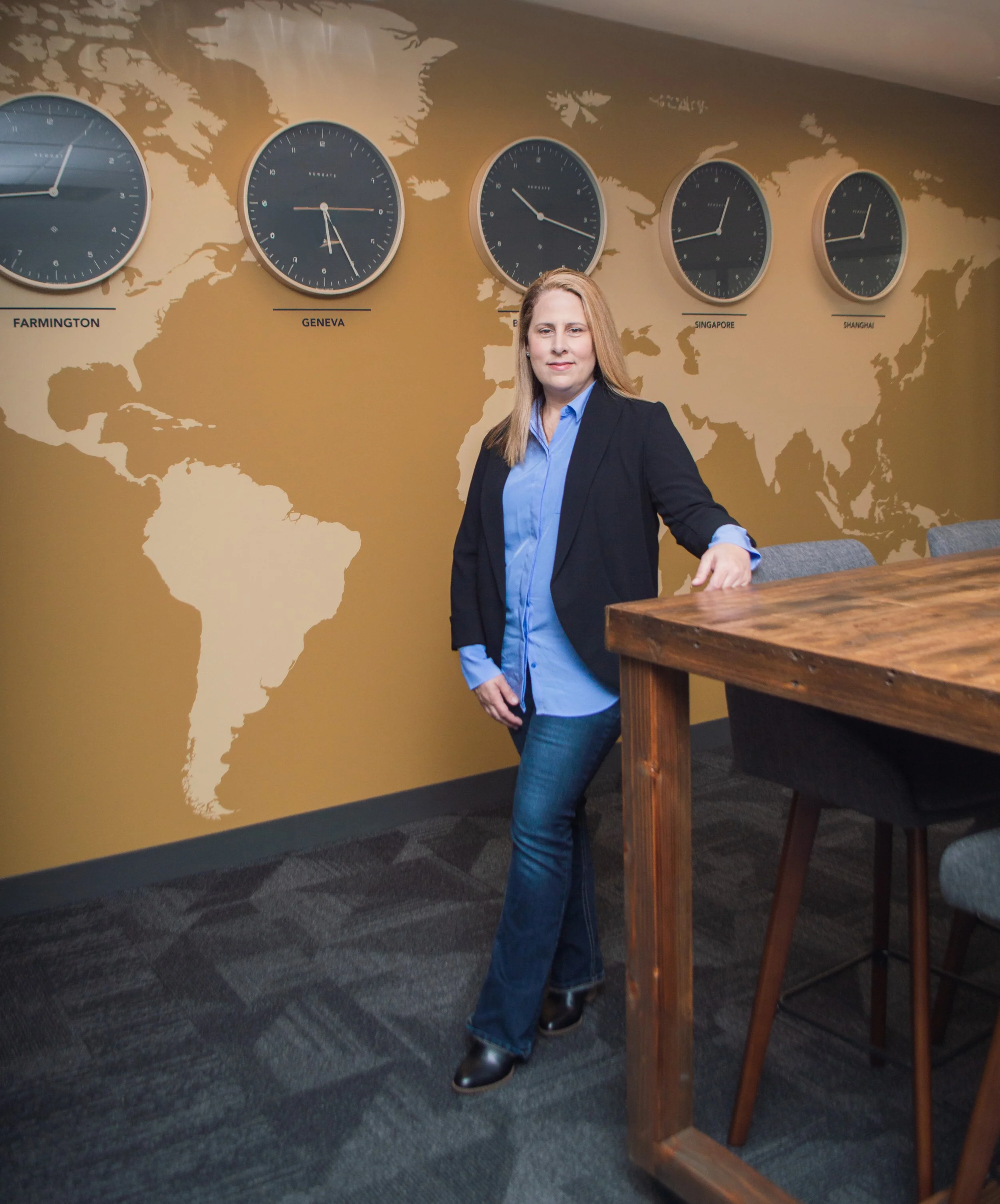 A woman in business attire standing in an office with a world map mural and five clocks showing different times labeled Farmington, Geneva, Singapore, Shanghai, and one partially visible, on the wall behind her.